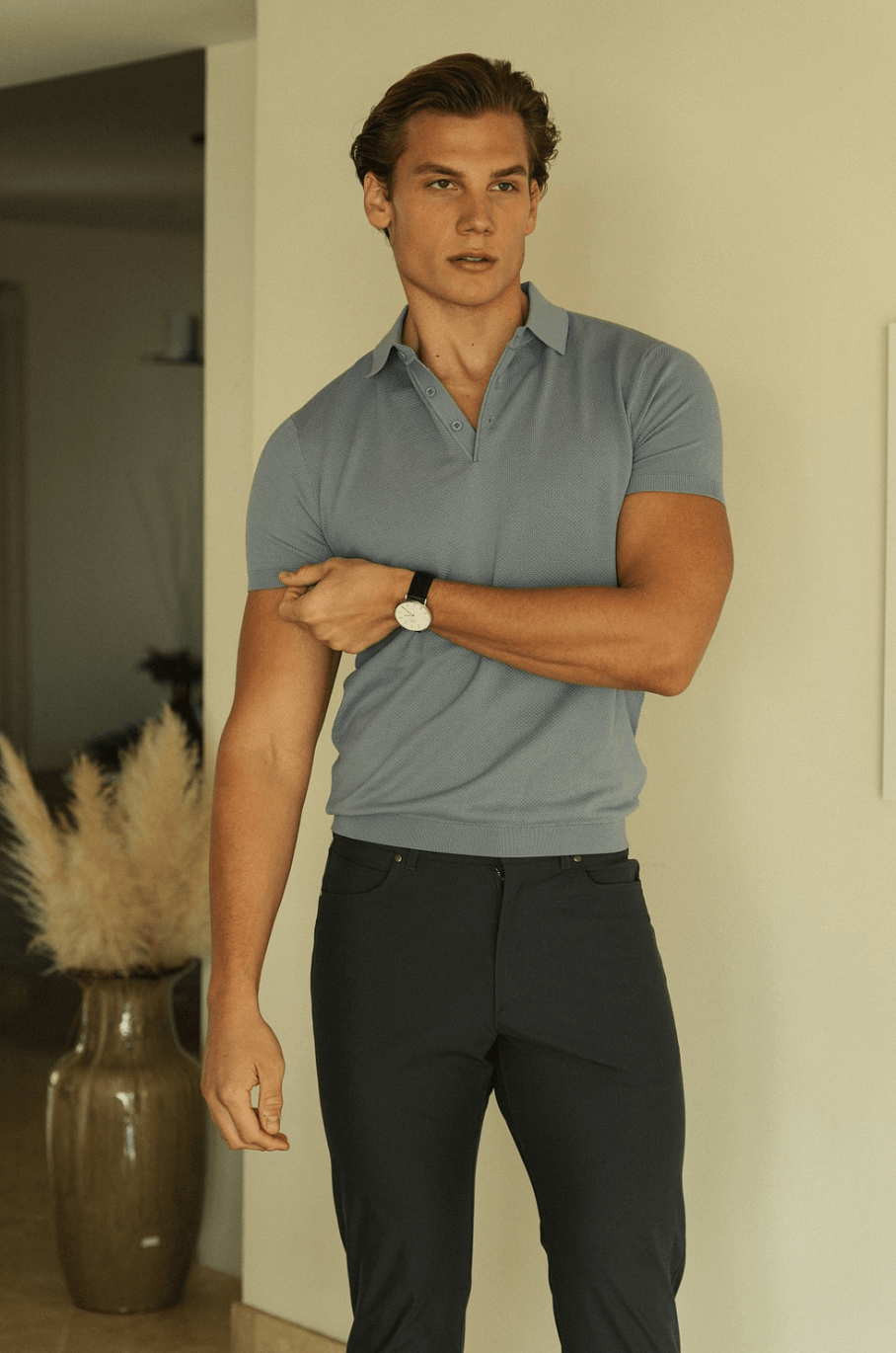 Young man with short hair wearing a light blue polo shirt and dark pants, standing indoors with a neutral expression.