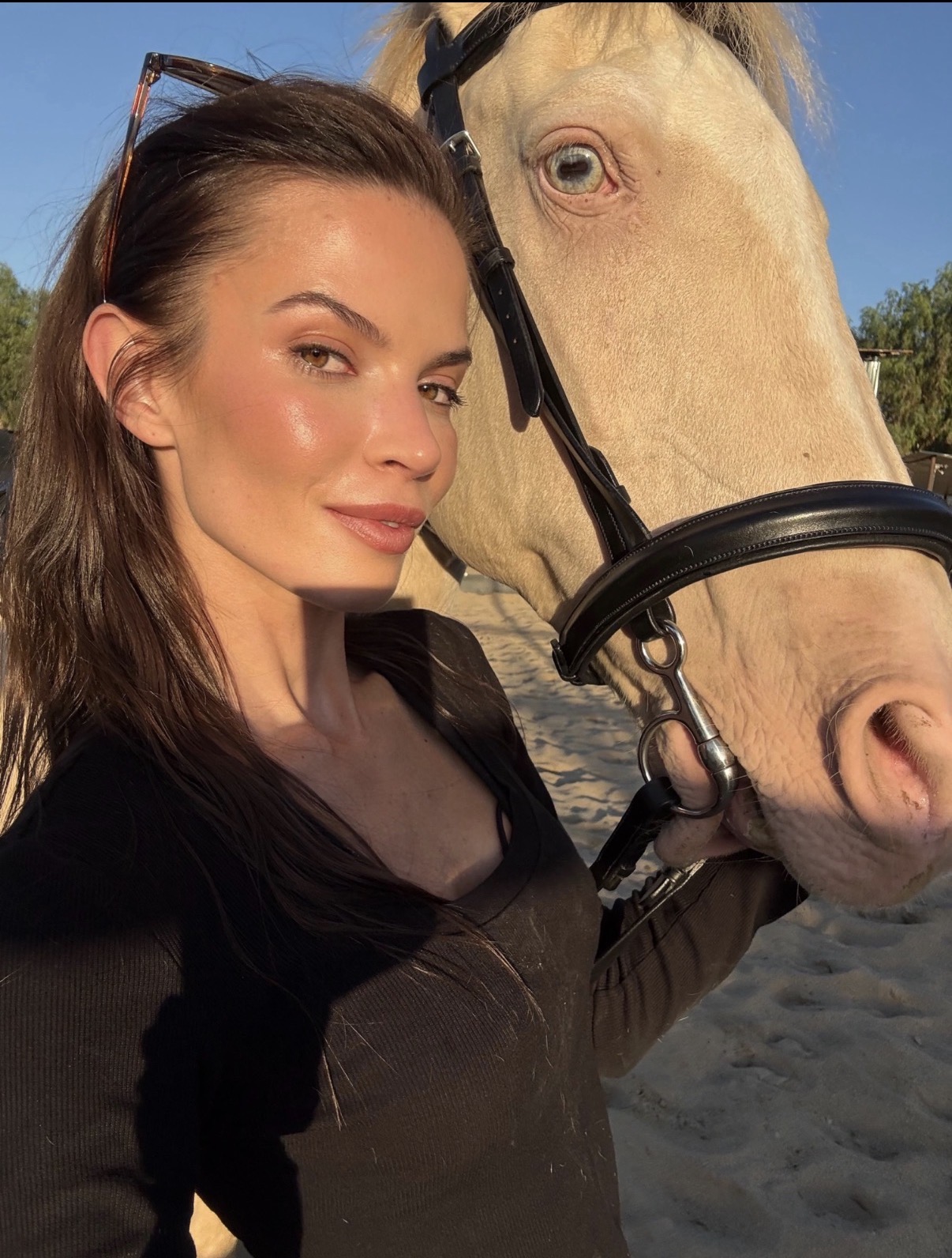 Woman with long brown hair and black top taking a selfie with a white horse outdoors during daytime.