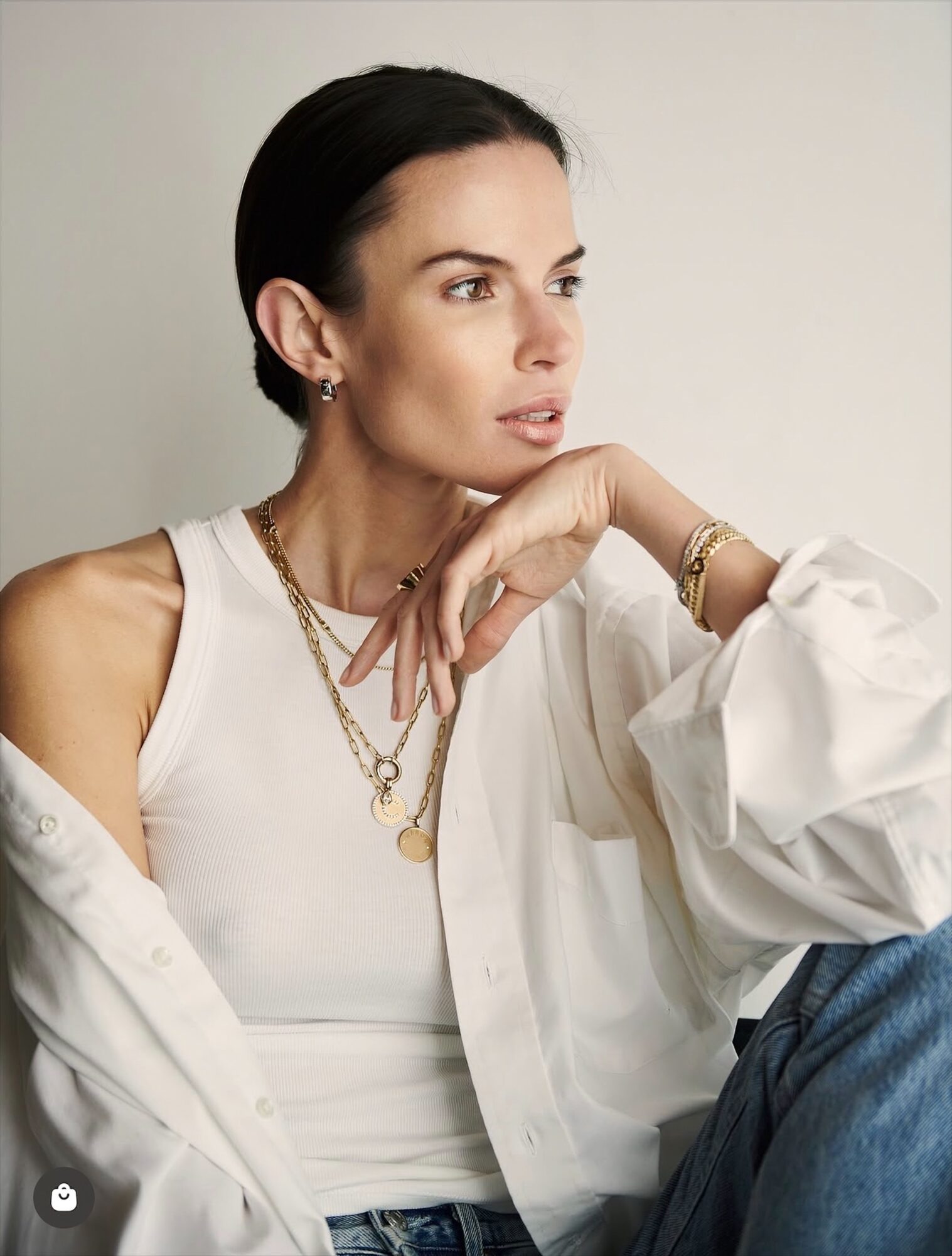Woman with dark hair styled back, wearing a white top and layered gold jewelry, sitting with her hand near her face.
