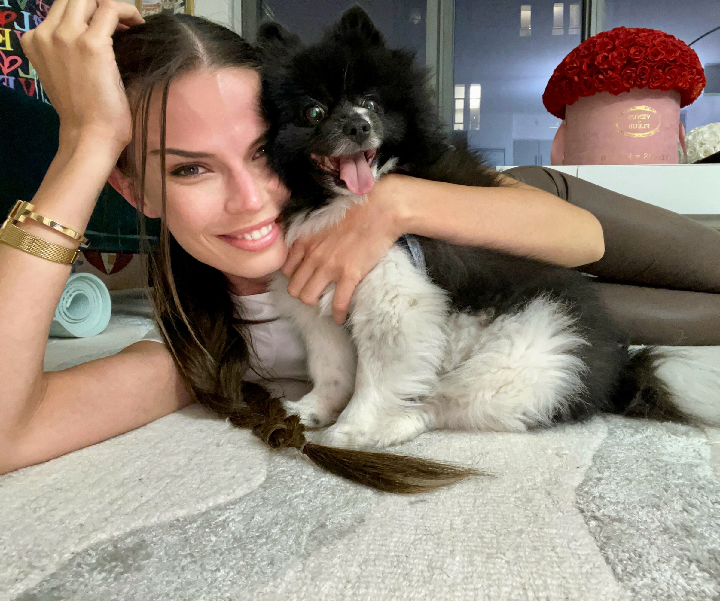 Woman lying on the floor with a black and white dog, smiling and hugging the dog, in a room with a window and a red hat on a shelf.