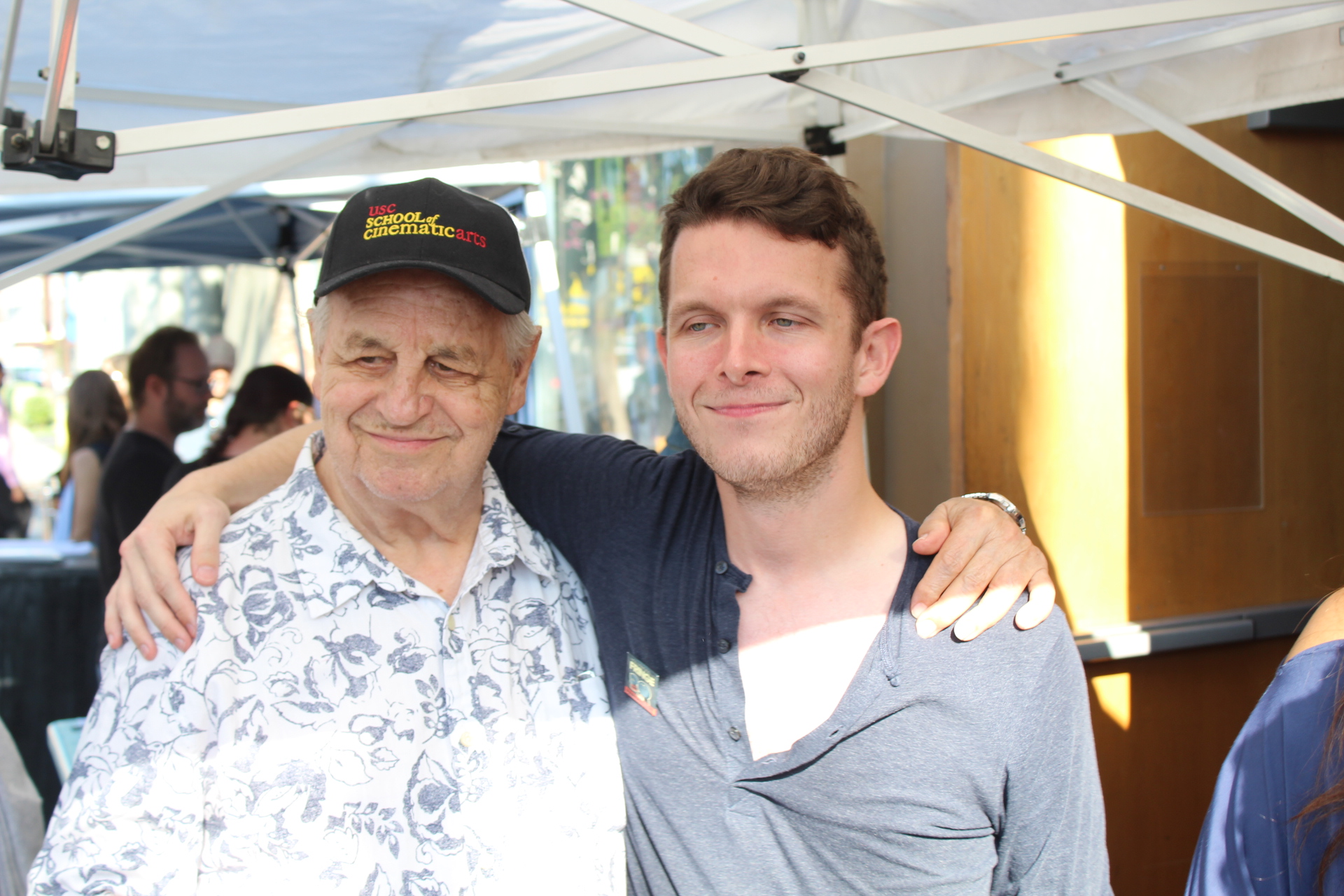 Two men smiling, one older with a cap and floral shirt, the other younger in a gray shirt, hugging under a tent.