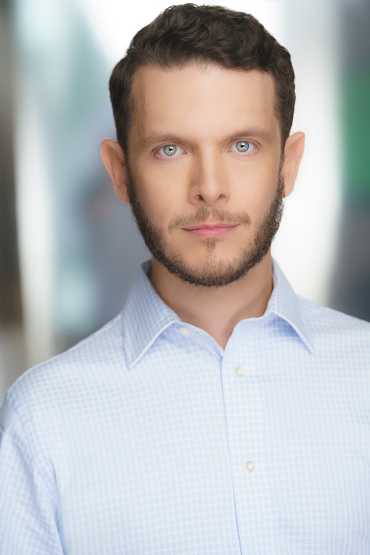 Portrait of a man with short dark hair, blue eyes, and a beard, wearing a light blue collared shirt, looking directly at the camera.