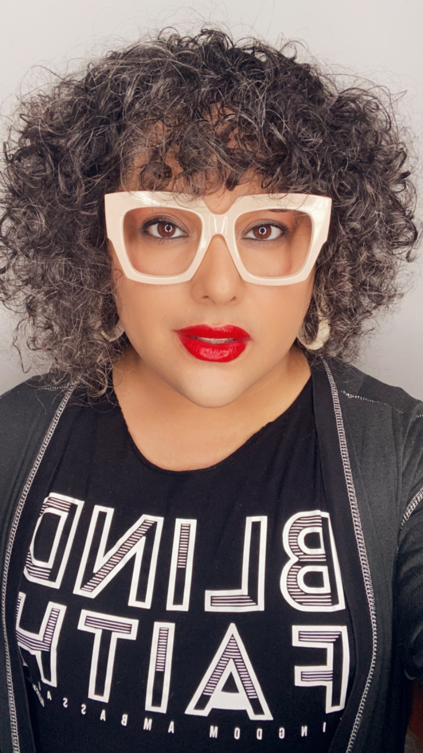 Woman with curly hair, white glasses, red lipstick, wearing a black shirt with white text, against a plain background.