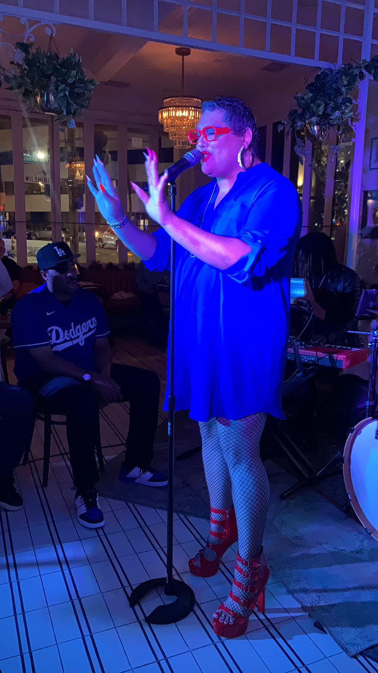 Person singing into microphone with hands raised, wearing glasses, a blue dress, fishnet stockings, and red high heels, in a decorated indoor setting.