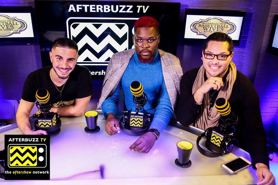 Three people sit at a table with microphones, cups, and a phone, in a studio with 'AFTERBUZZ TV' signs behind them.