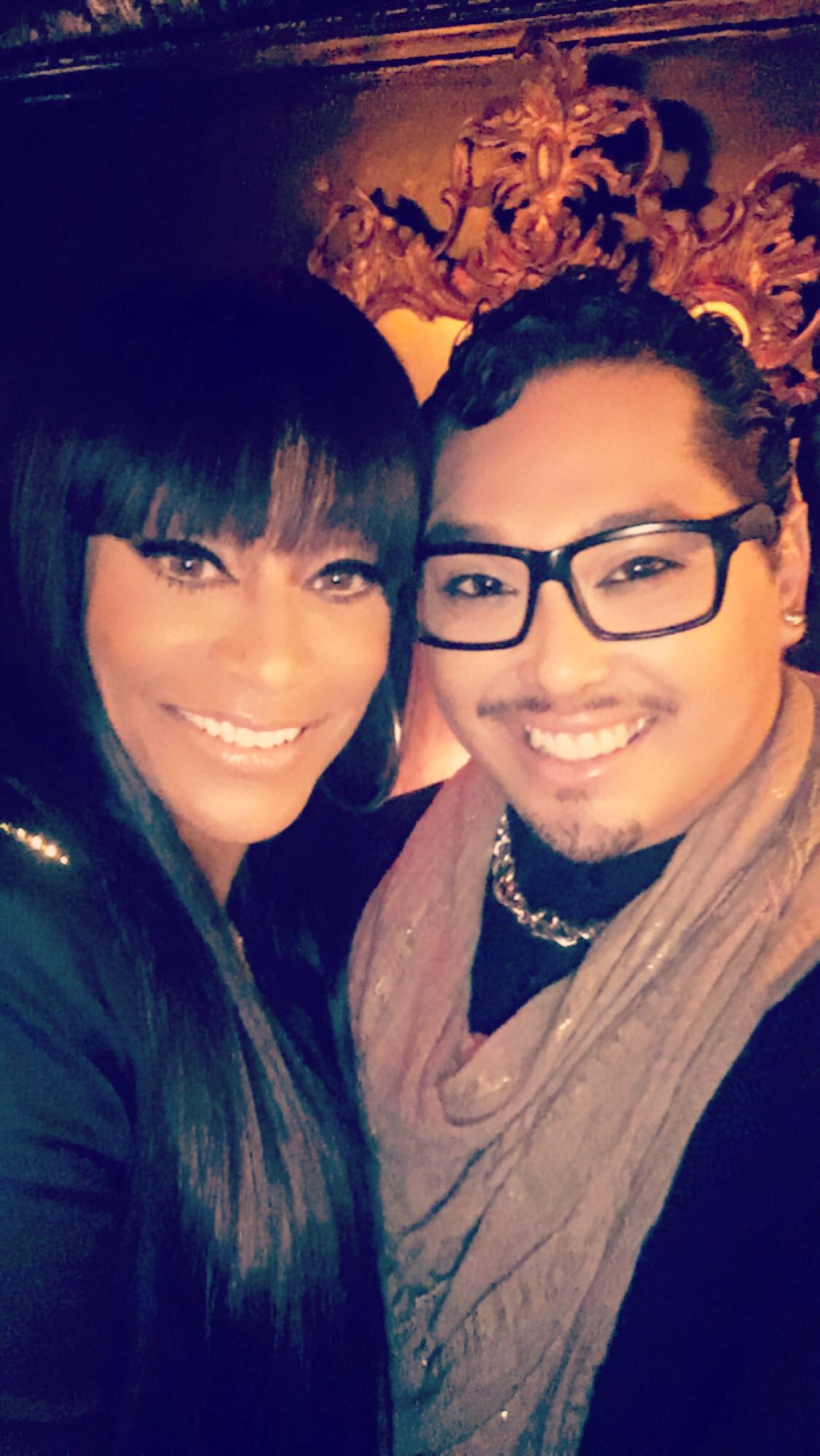 Two people smiling closely, one with long dark hair and the other with glasses and short hair, in a dimly lit setting.