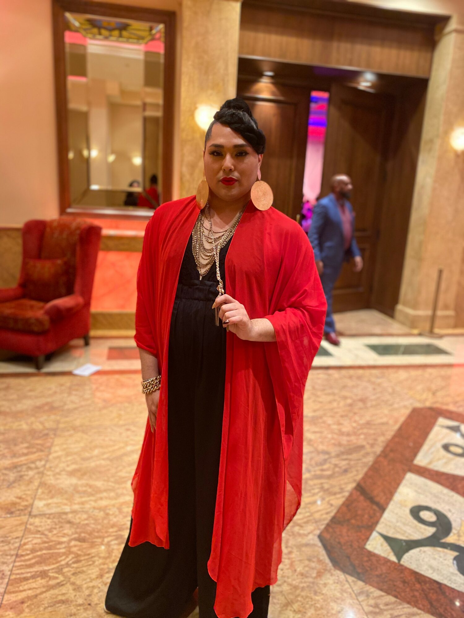Woman in red coat and black dress standing in a hotel lobby with wooden walls and a red armchair.