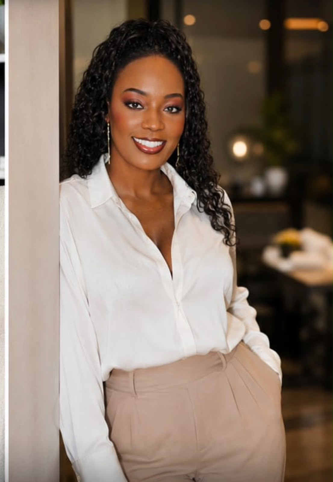 Woman with curly hair smiling, wearing a white blouse and beige pants, standing indoors near a wall.