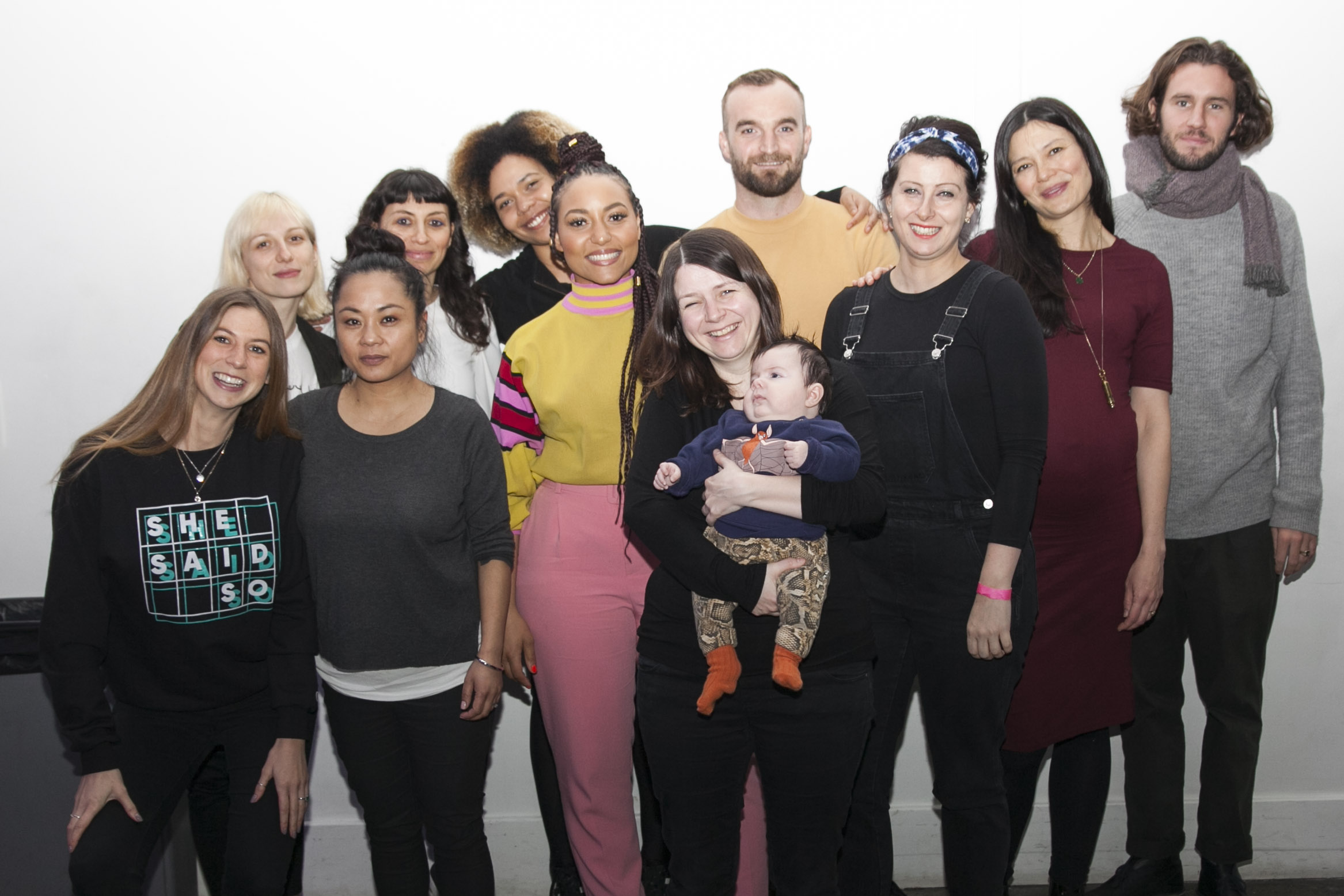 Group of diverse people standing together, smiling, with a baby in the center, against a plain white background.