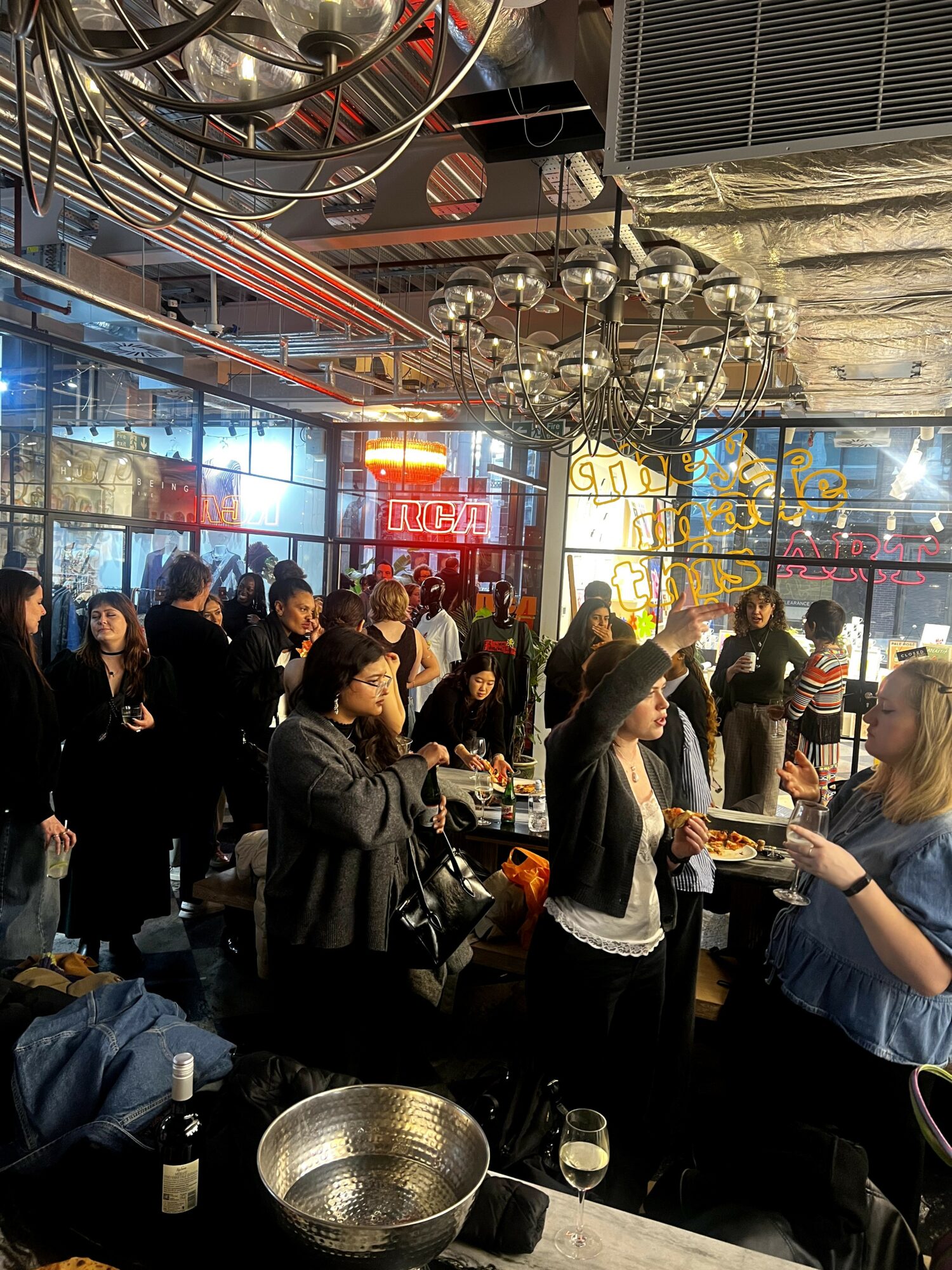 People gather in a modern indoor space with large windows, hanging lights, and a bar area, engaging in conversations and activities.