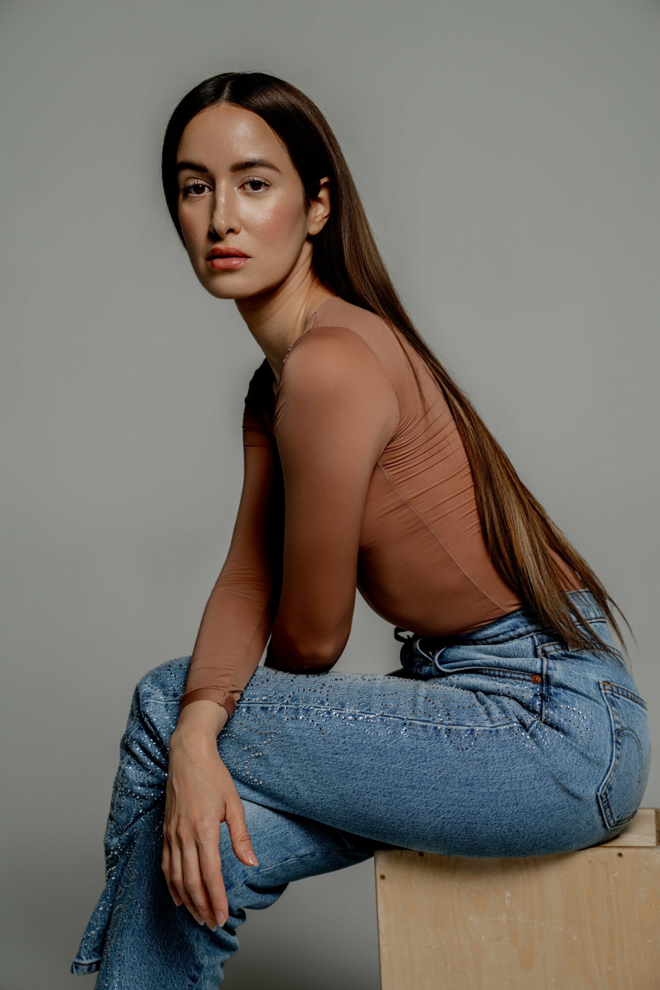 Young woman with long hair sitting on a wooden block, wearing a sheer top and jeans, against a plain background.