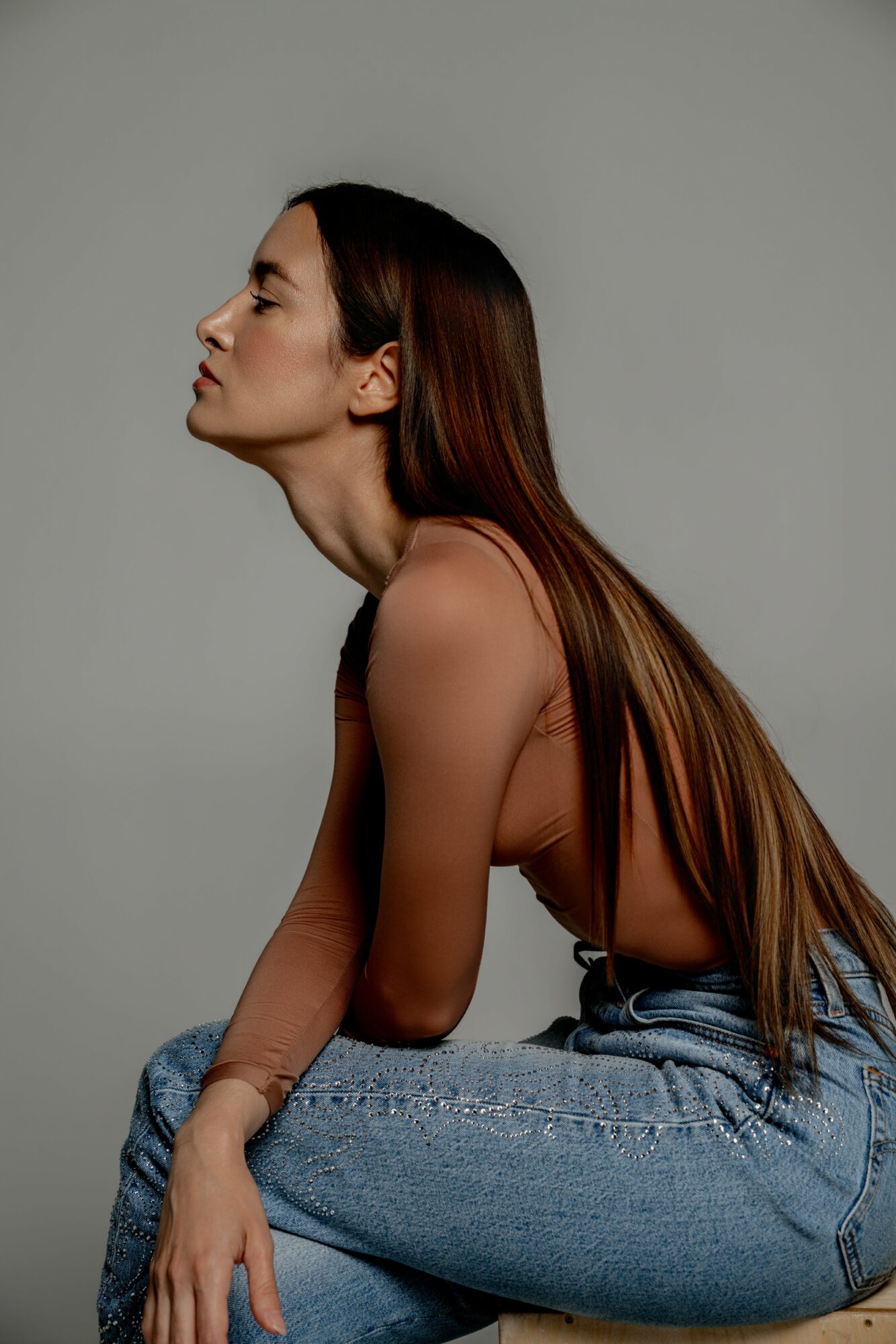 Side profile of a woman with long hair, sitting with her arms resting on her knees, against a plain background.