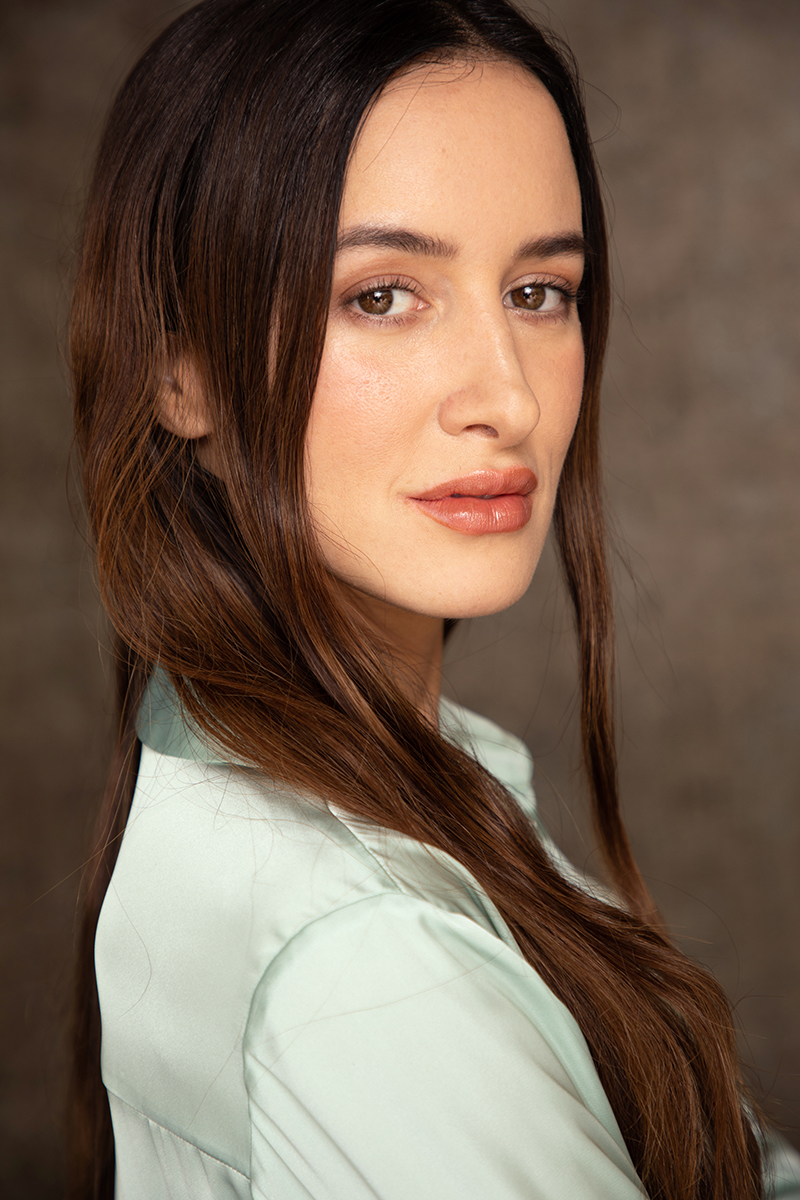 Young woman with long brown hair and light skin, looking at the camera, neutral expression, plain background.