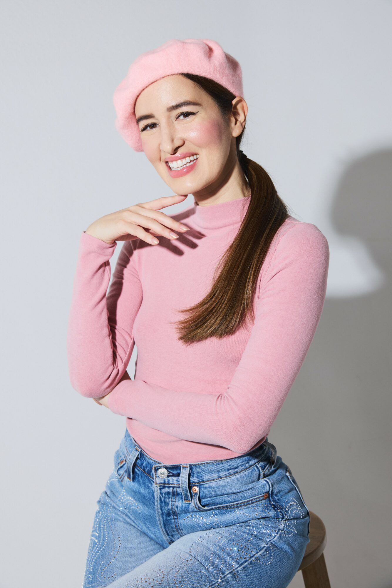 Young woman smiling, wearing pink beret and sweater, with long brown hair, against a light background.