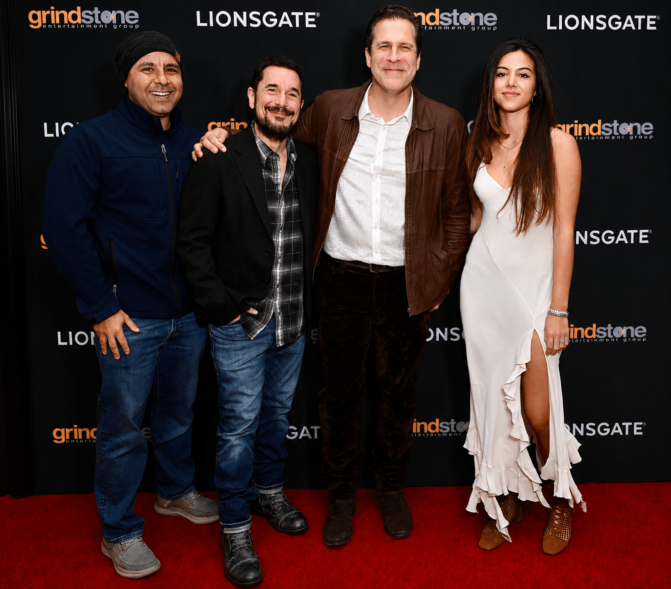 Four people standing on a red carpet in front of a black backdrop with logos, smiling and posing for the photo.