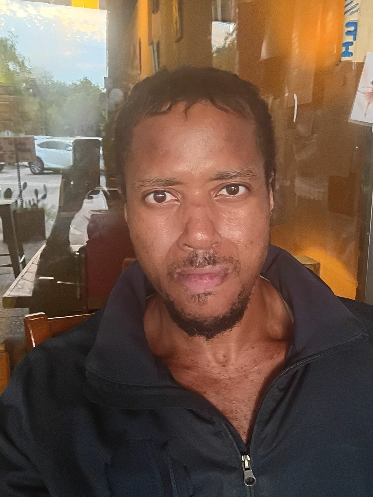 Man with short dark hair and a goatee, sitting indoors near a window with sunlight, looking at the camera.