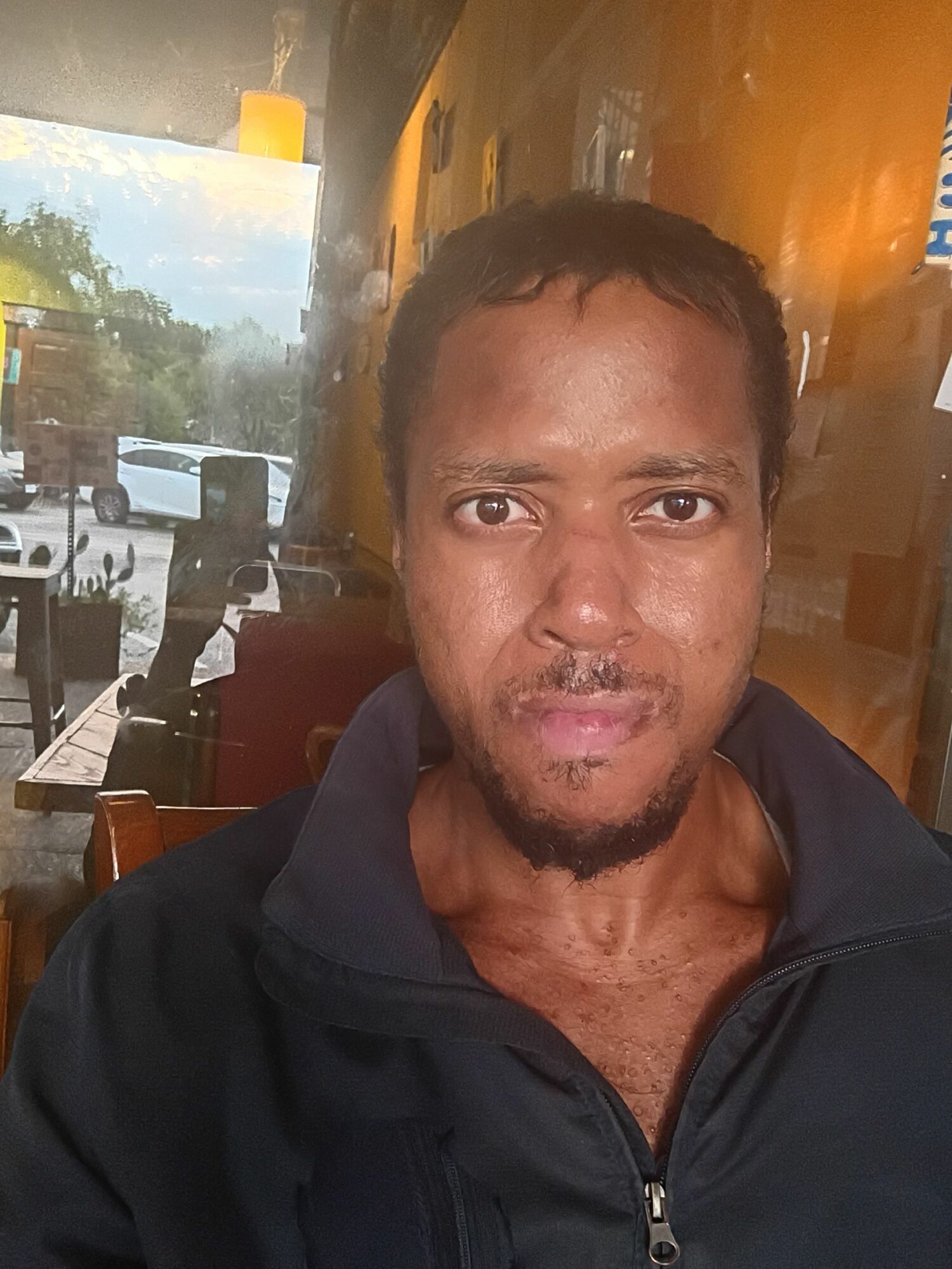 Man with short dark hair and facial hair, indoors with a window showing outdoor scenery, wearing a dark jacket.