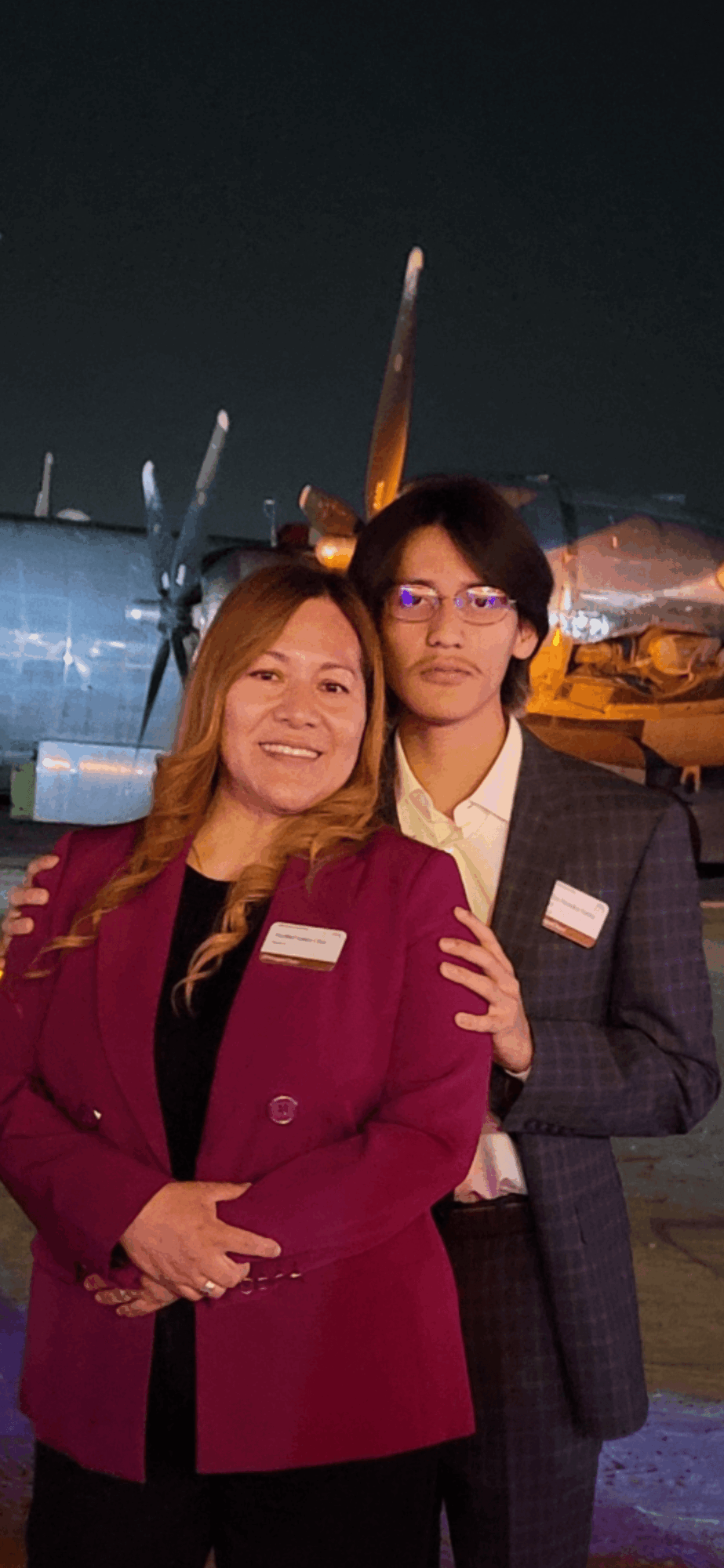 Two women standing outdoors at night, smiling, with aircraft in the background, one wearing glasses and a maroon blazer.