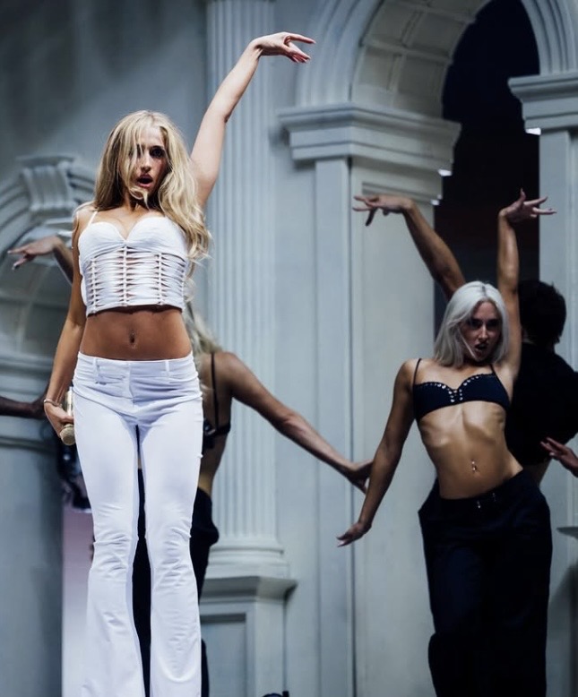 Two women dance in a grand hall with white columns, one in white pants and a crop top, the other in black pants and a crop top.