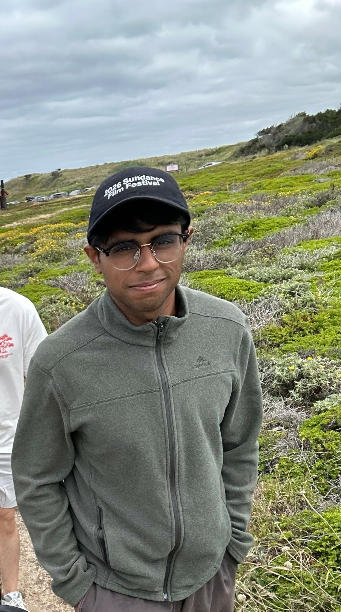 Young person wearing glasses and a cap outdoors in a grassy landscape with cloudy sky.