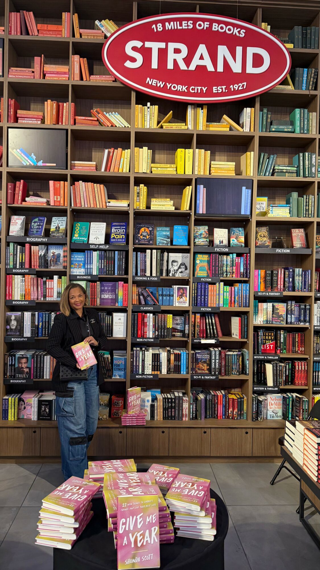 Person standing in front of tall bookshelf filled with colorful books, holding a book, with a sign reading 'Strand' above.