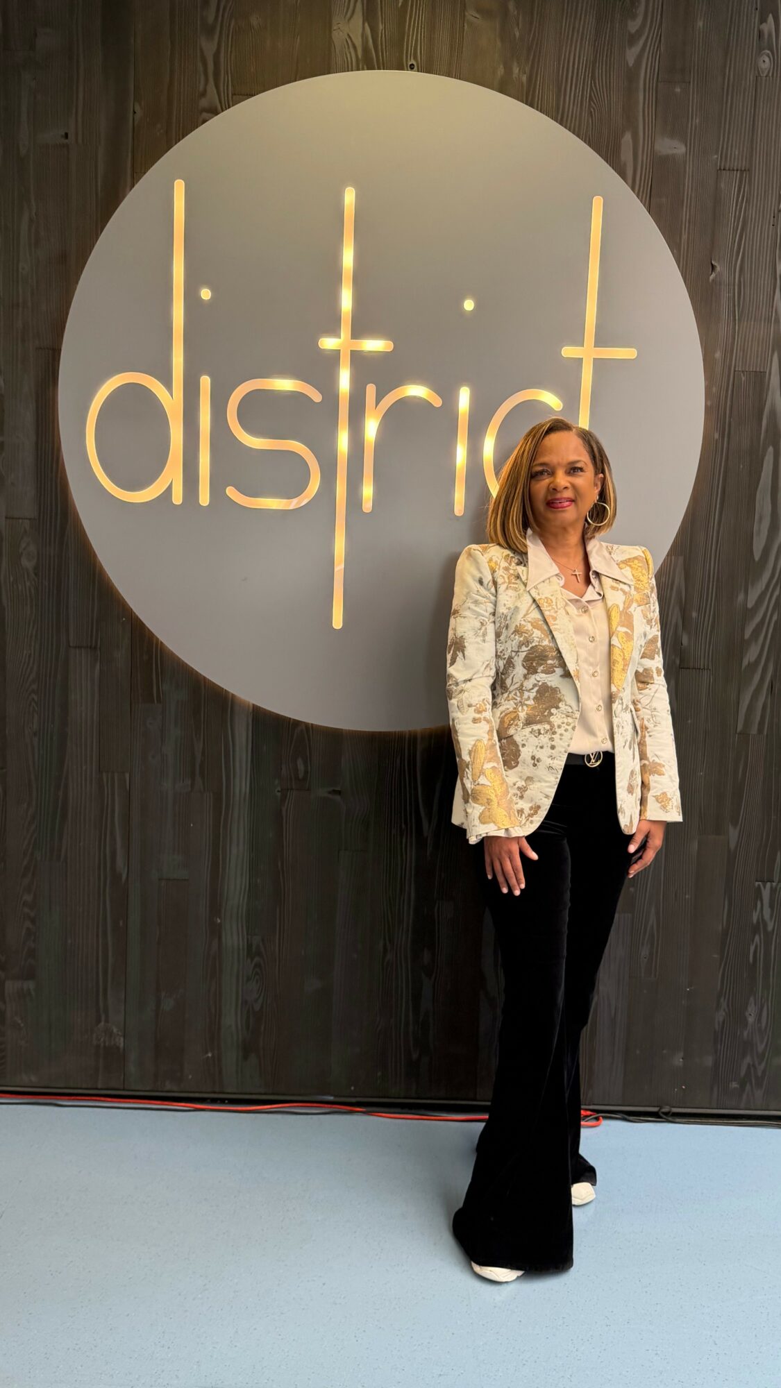 Woman standing in front of a large circular sign with the word 'district' in gold letters, wooden wall background.