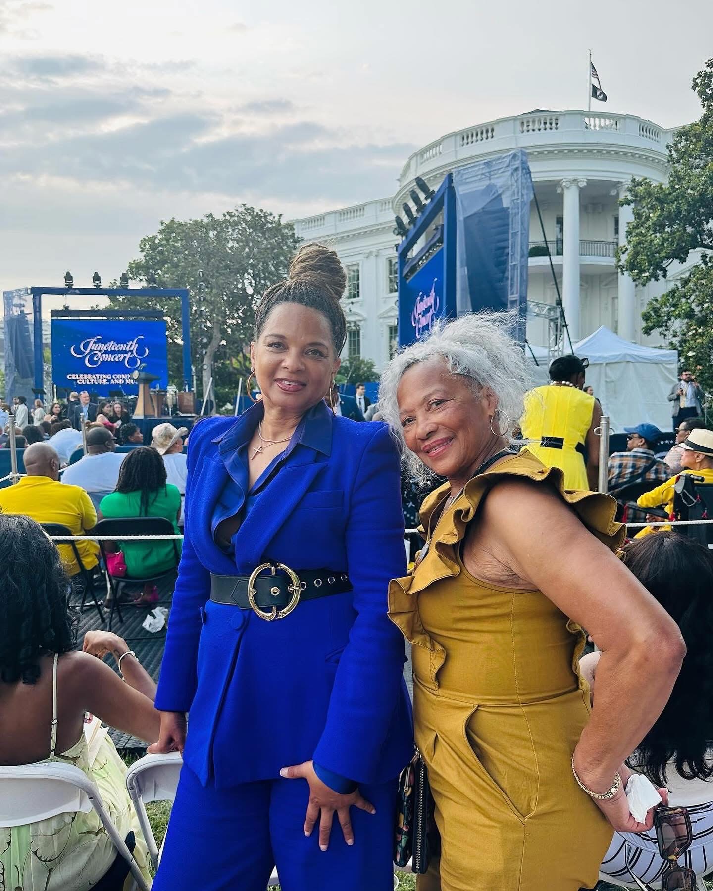 Two women smiling outdoors in front of the White House, one in a blue outfit and the other in a yellow dress, with a crowd and screens in the background.