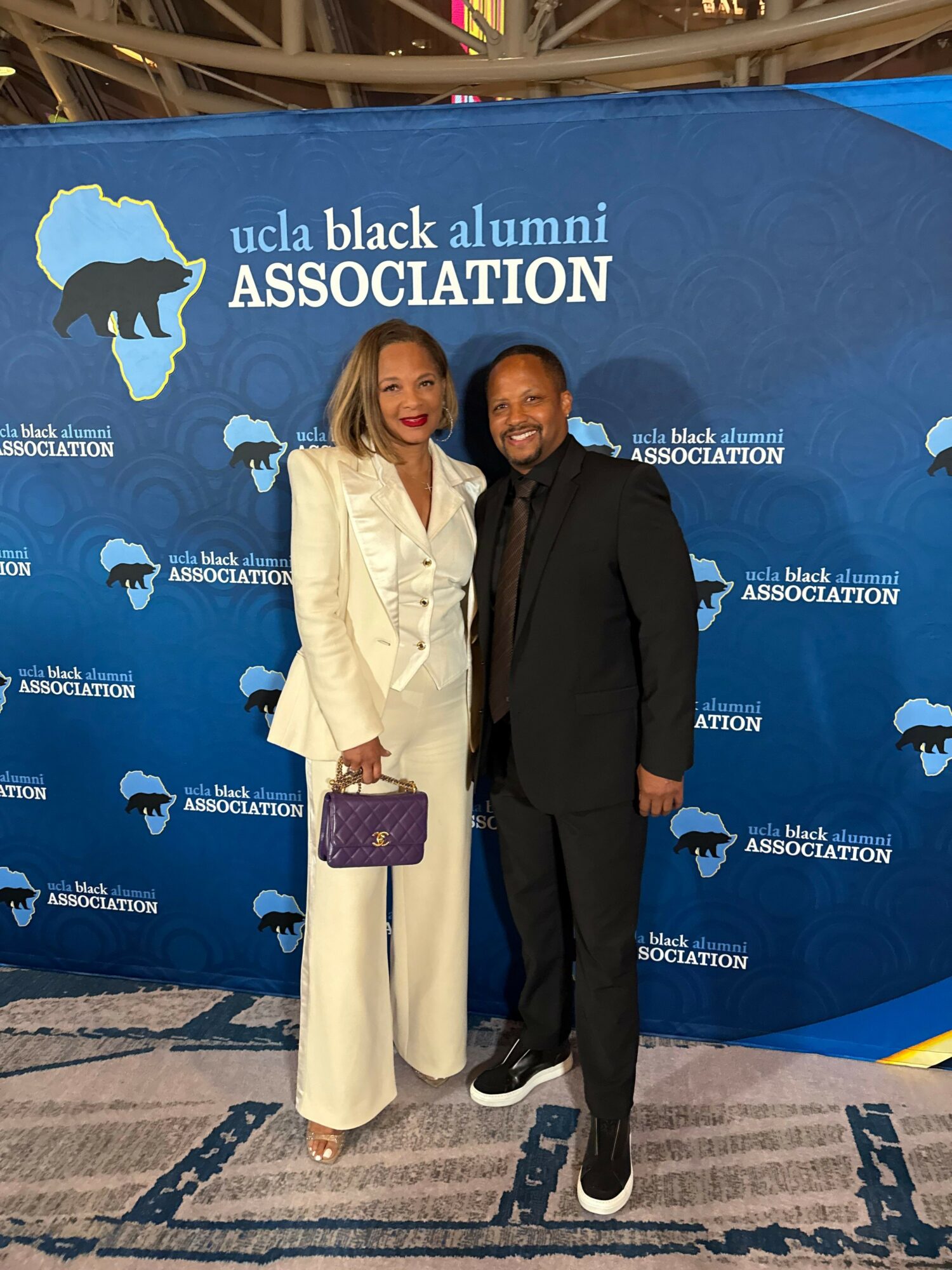 Two women standing on a blue carpet with a backdrop displaying the UC Black Alumni Association logo, one in a black suit and the other in a cream-colored suit holding a purple clutch.