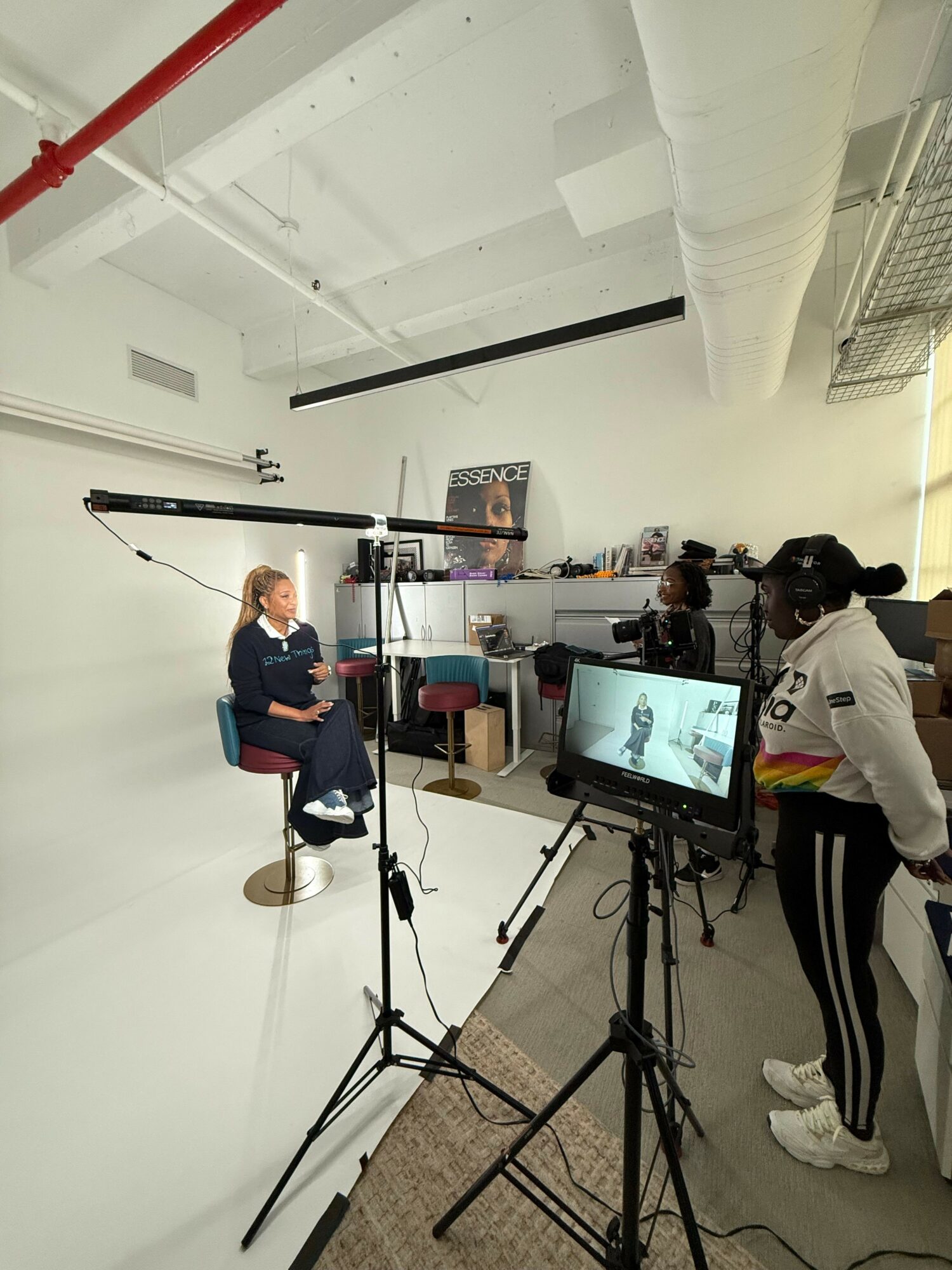 Person sitting on a stool in a studio with camera equipment and a monitor, recording or streaming content.