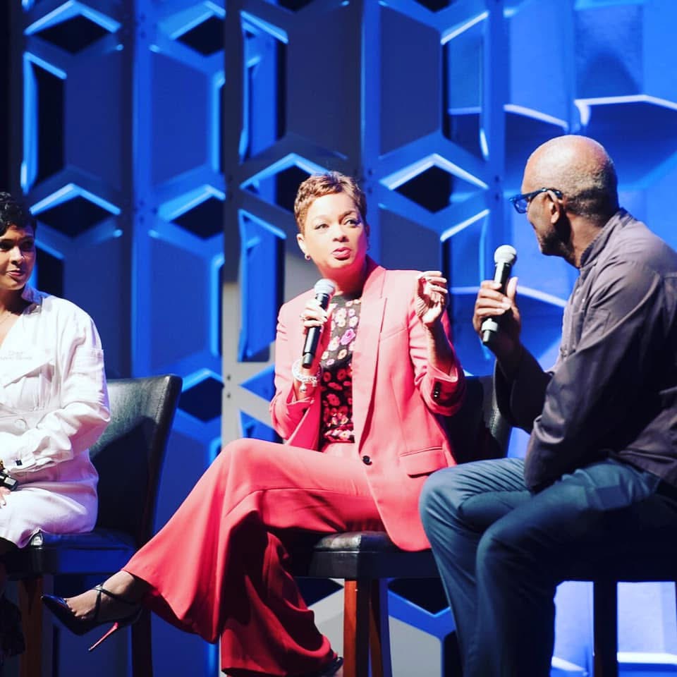 Three people sitting on stage with a blue geometric background, two holding microphones, engaged in conversation.