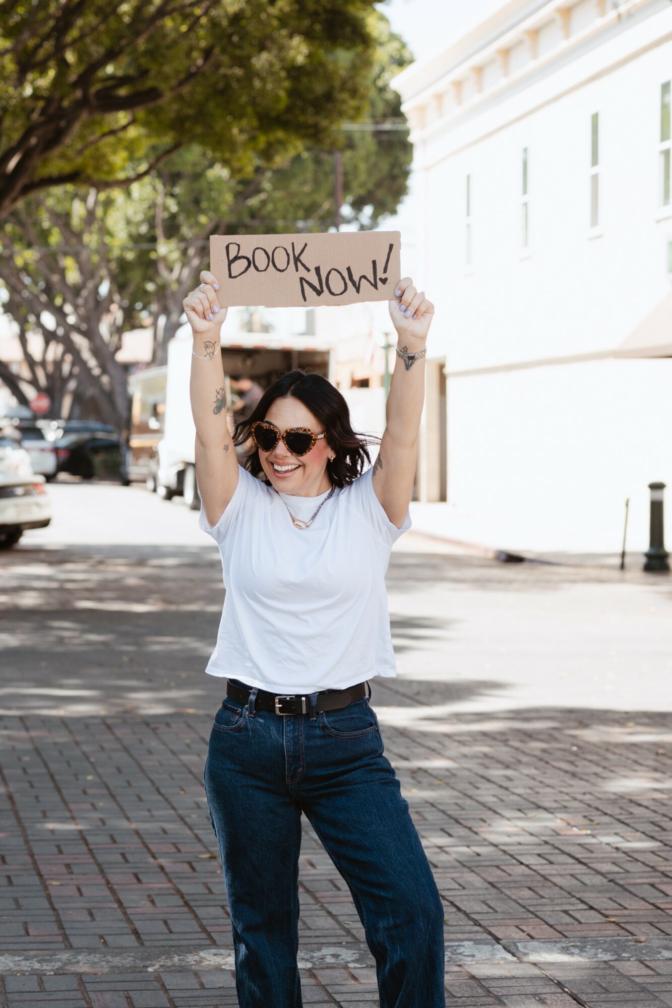 Woman holding a sign that says 'Book Now!' smiling outdoors on a sunny day.