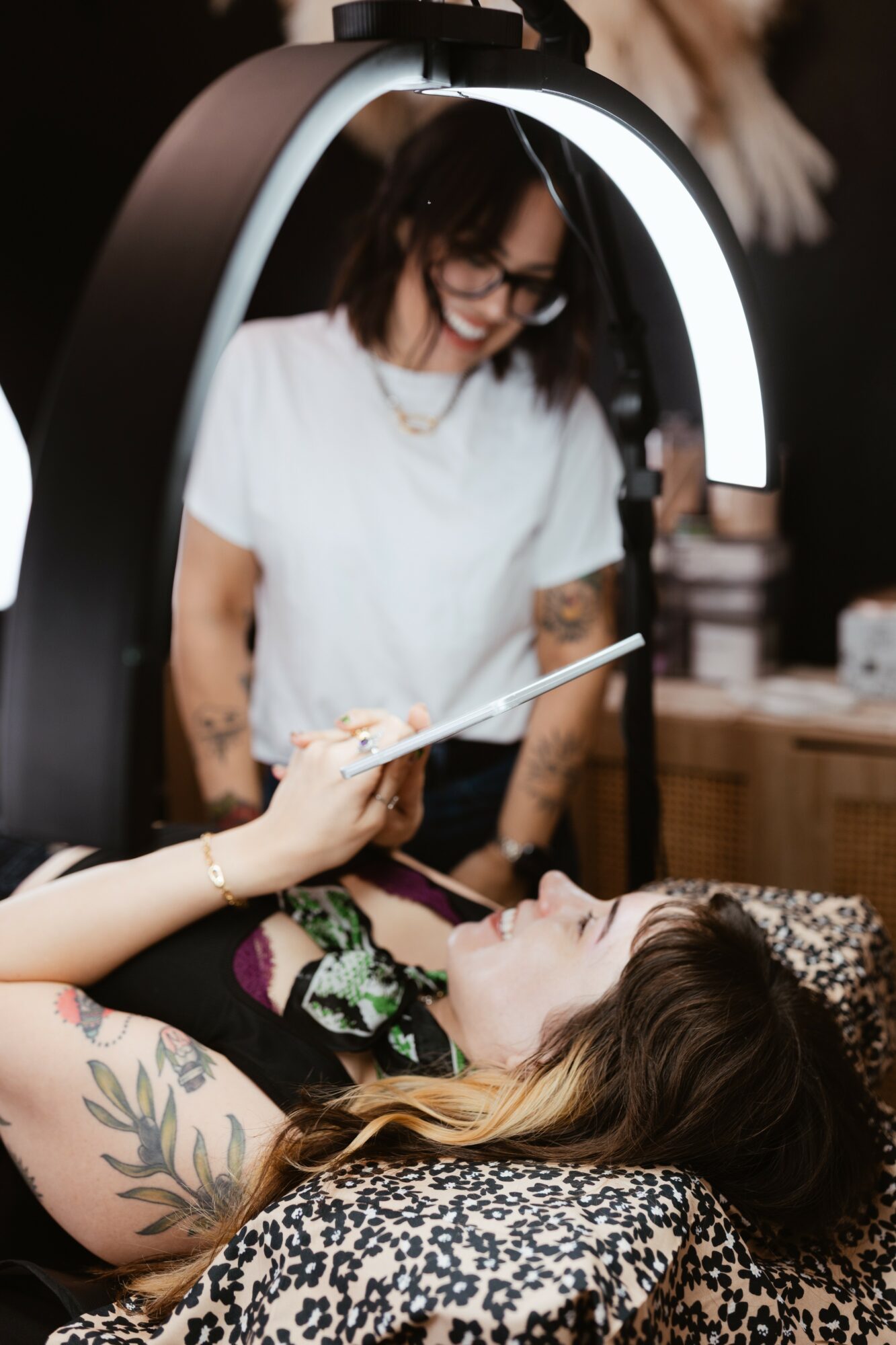 A woman lying on a bed with floral sheets, receiving a facial treatment, while a professional applies a mask or cream.