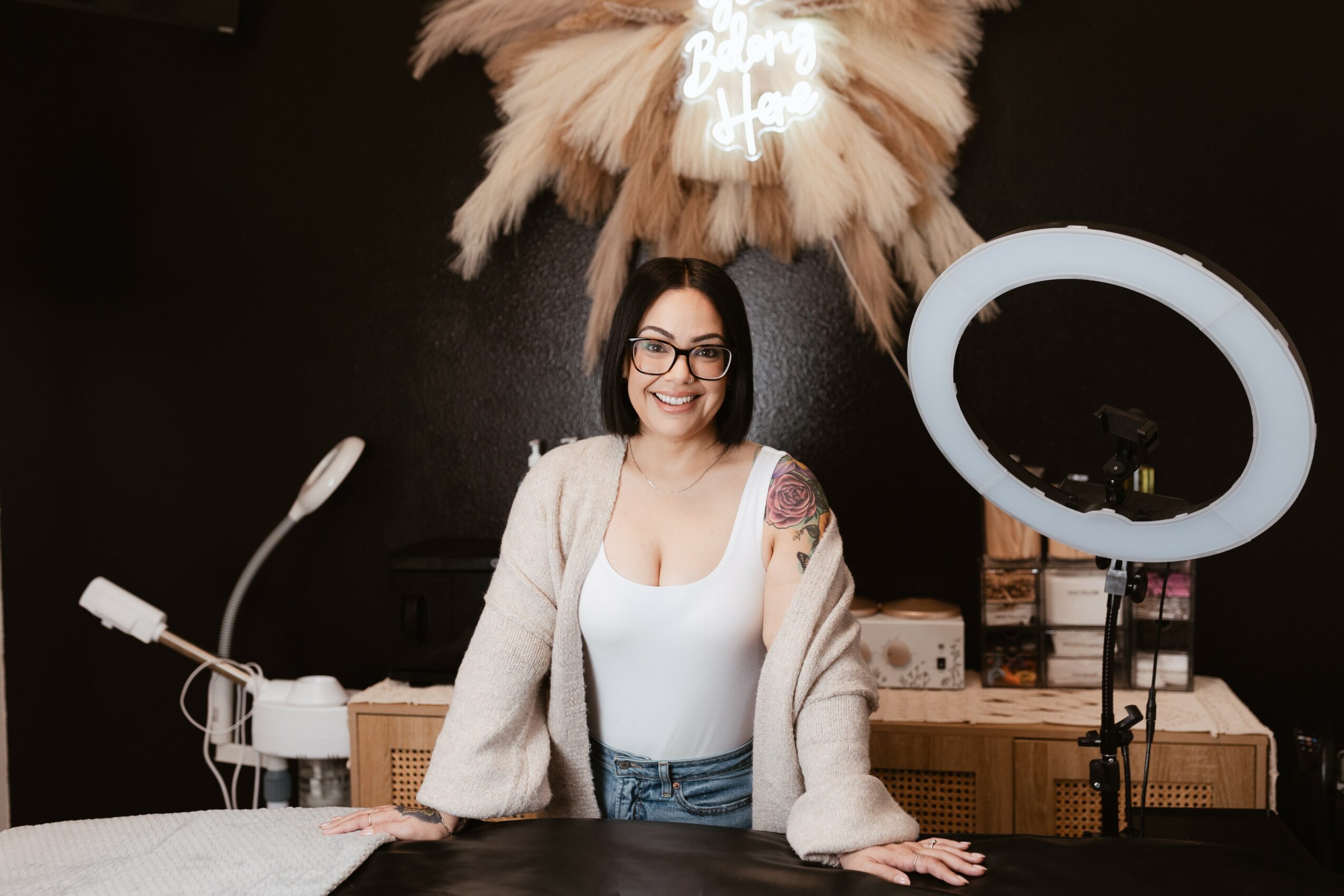 Woman with glasses and dark hair smiling, standing in front of a black wall with a ring light and fluffy decoration above her head.