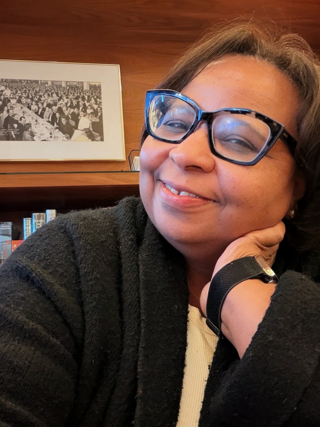 Woman with glasses smiling, resting her chin on her hand, in a cozy indoor setting with a framed photo on the wall behind her.