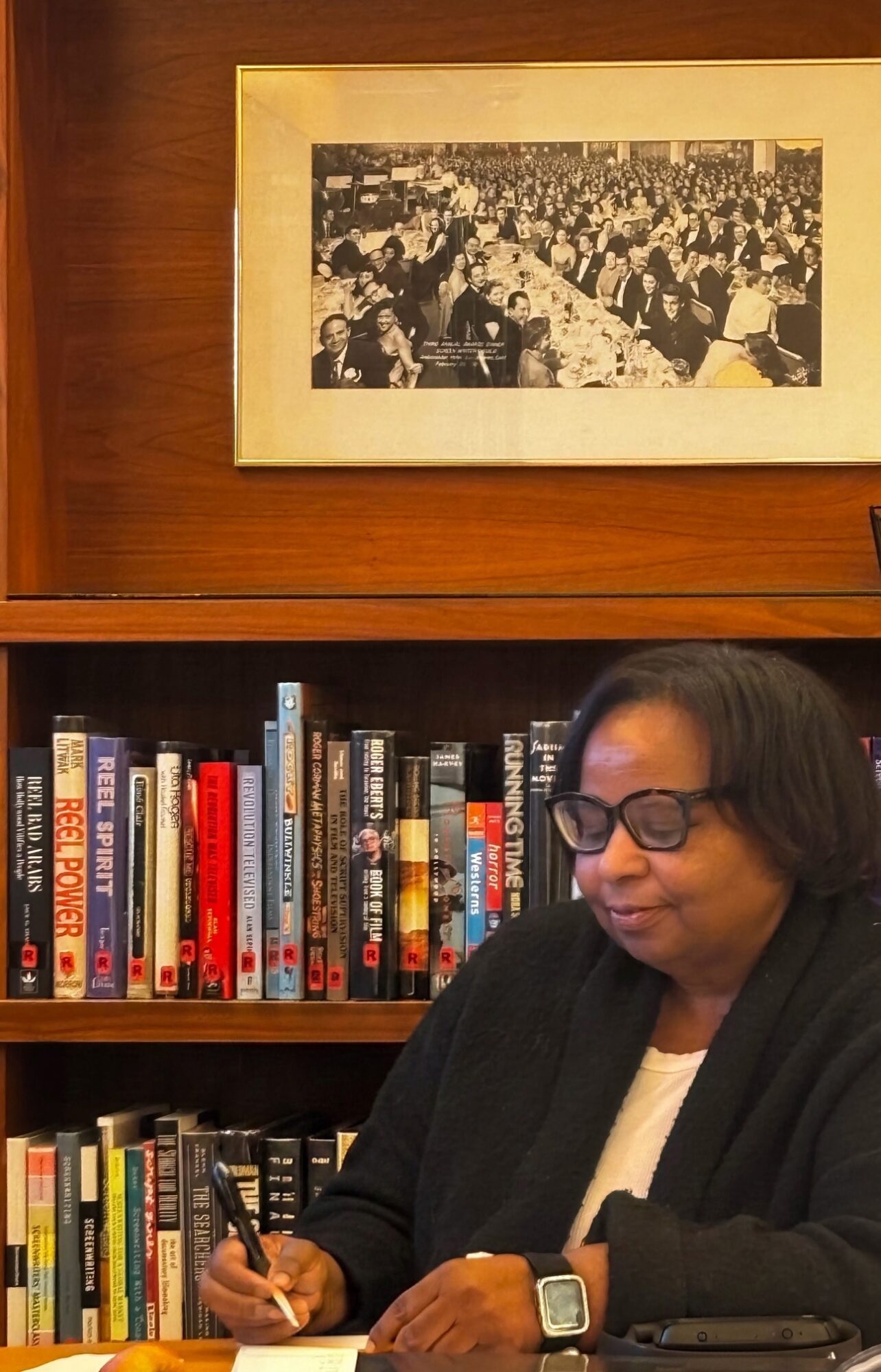 Woman with glasses and a smartwatch sitting at a table, holding a pen, in front of a bookshelf with a framed photo above.