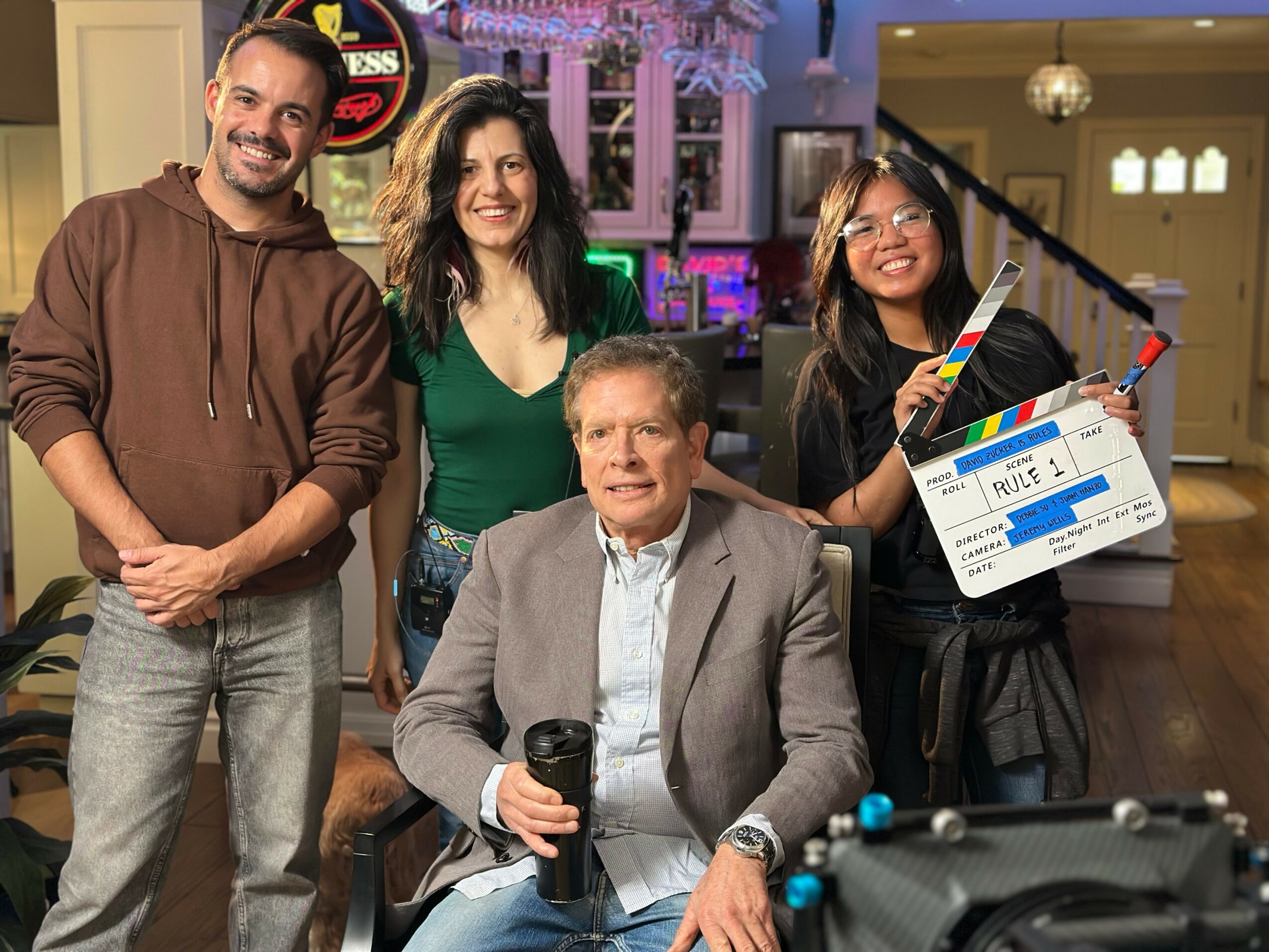 Four people in a room, one sitting at a table with a camera, others standing behind, smiling, with a clapperboard, colorful background.