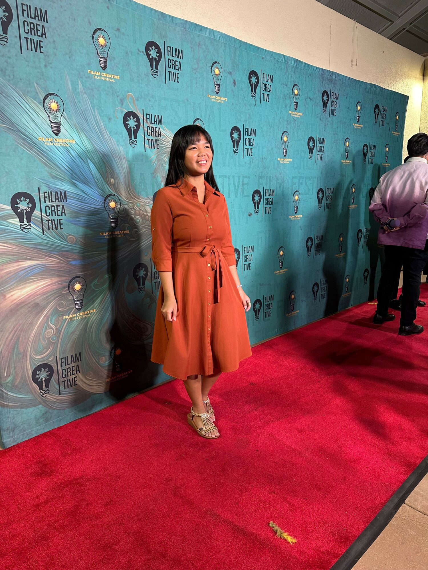 Woman in orange dress standing on red carpet with blue backdrop behind her, event setting, people in background.