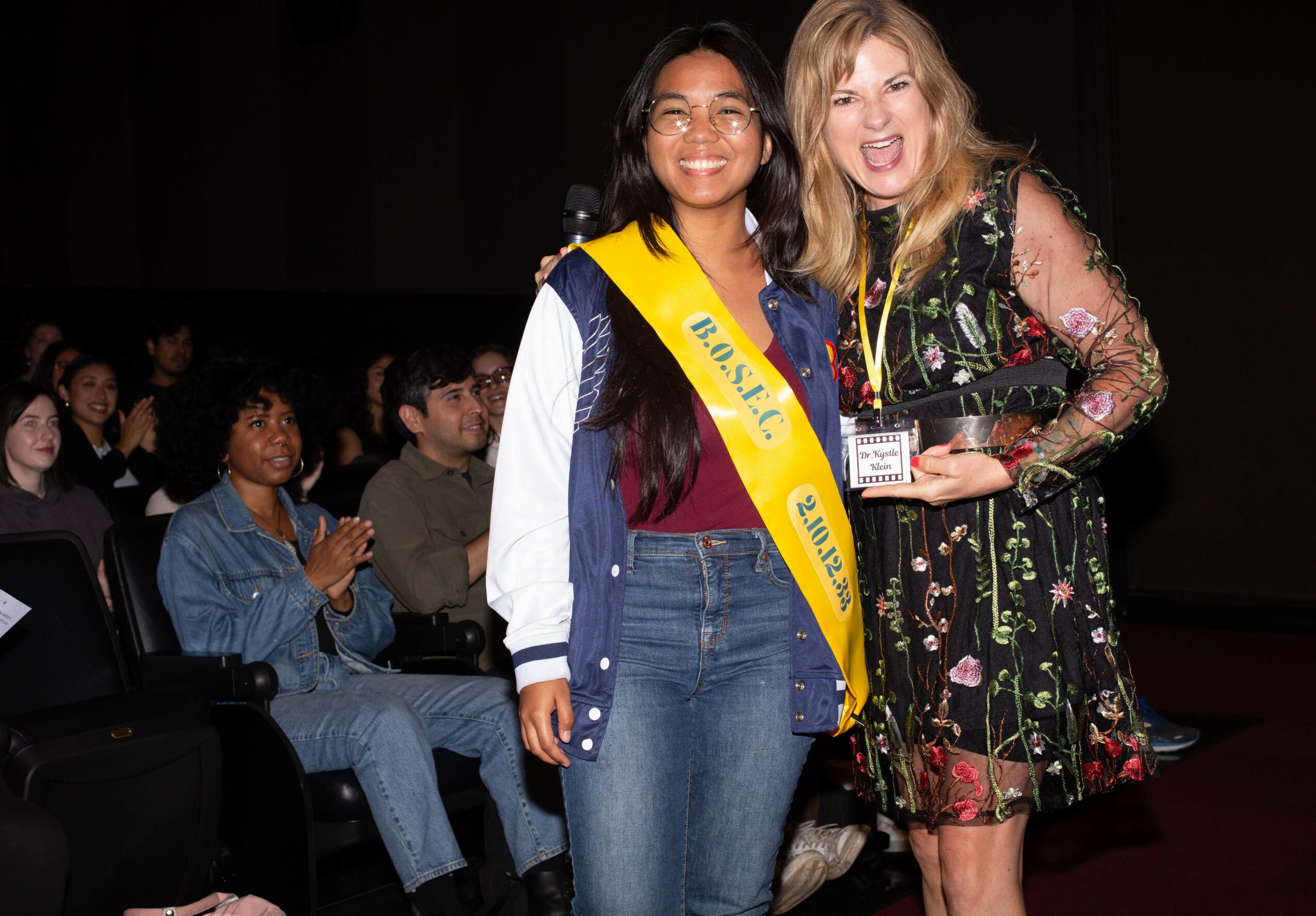 Two women smiling, one wearing a sash, at an indoor event with seated audience in background.