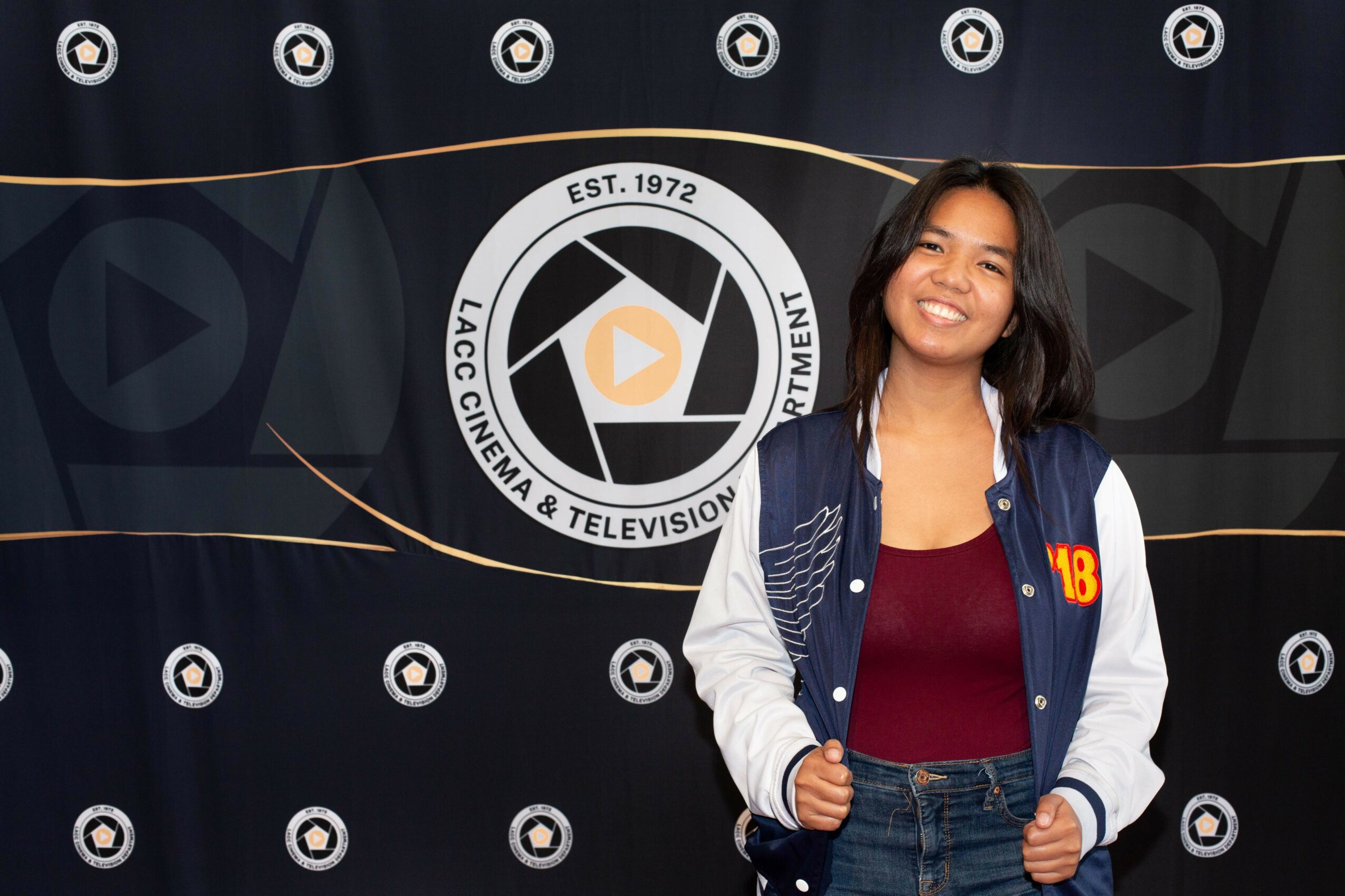 Young woman smiling, standing in front of a backdrop with a logo, wearing a blue and white jacket and a maroon top.