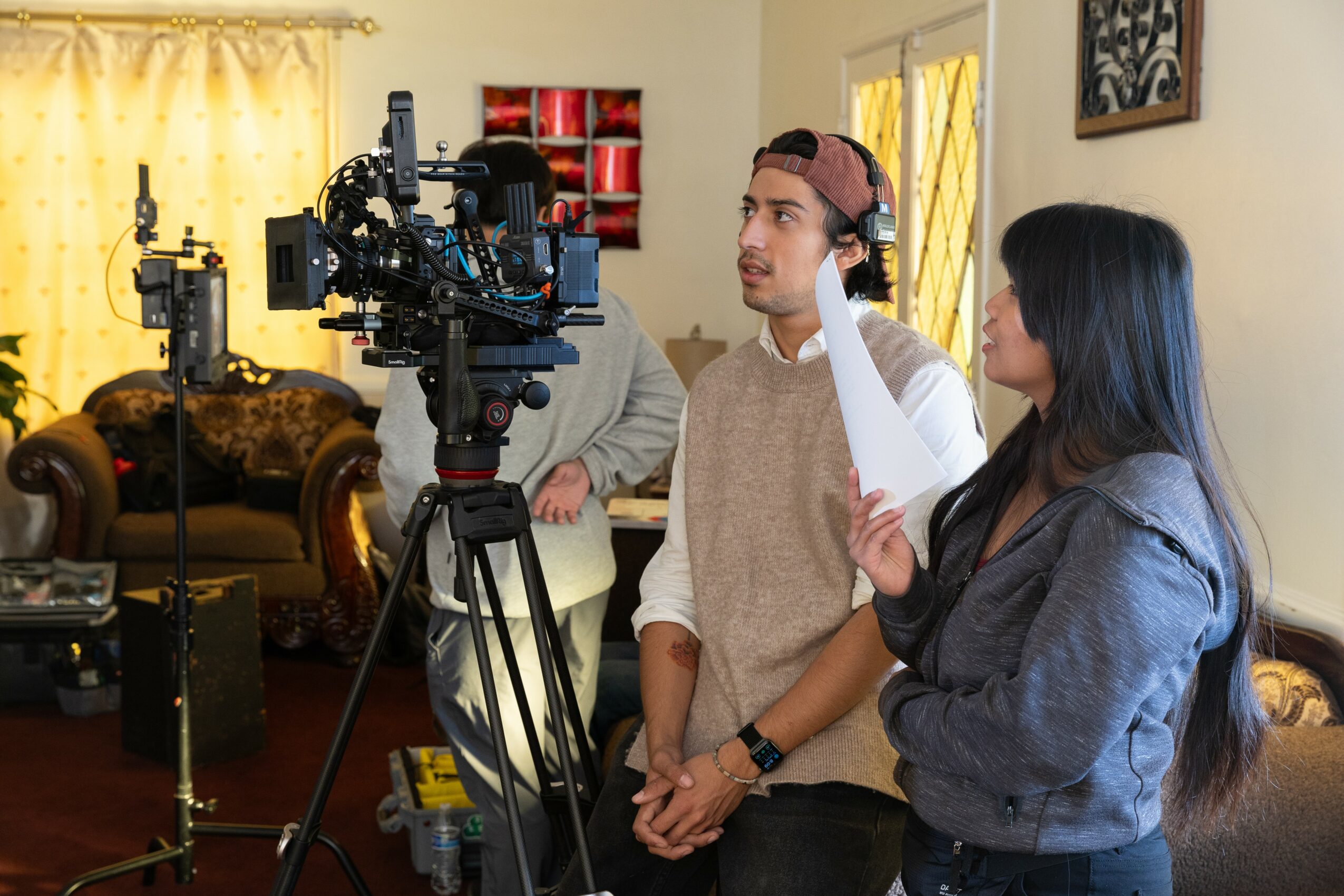 Two people on a film set, one with headphones and the other speaking, with camera equipment in a living room.