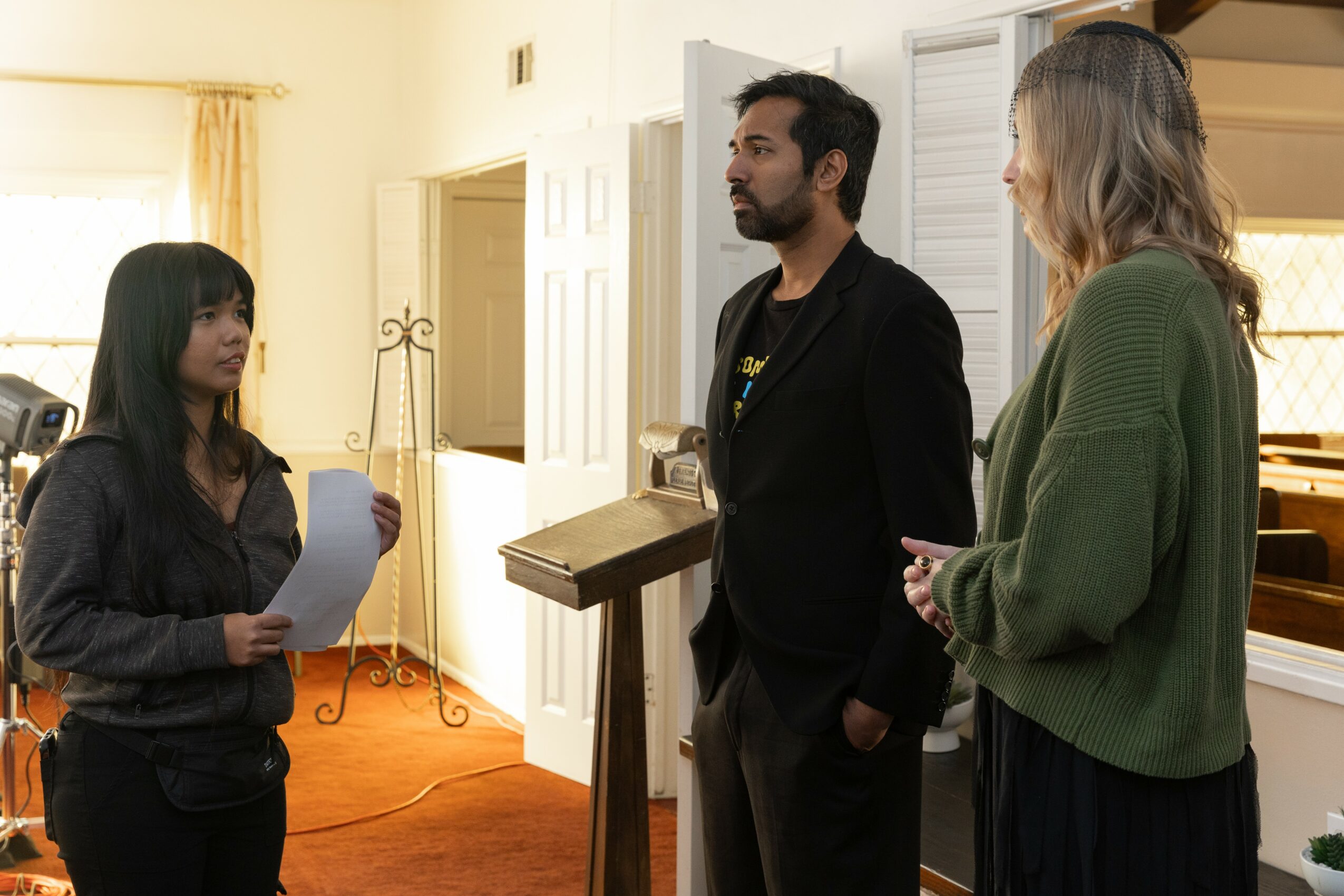 Three people standing and talking indoors, one holding papers, in a room with yellow walls and a window.