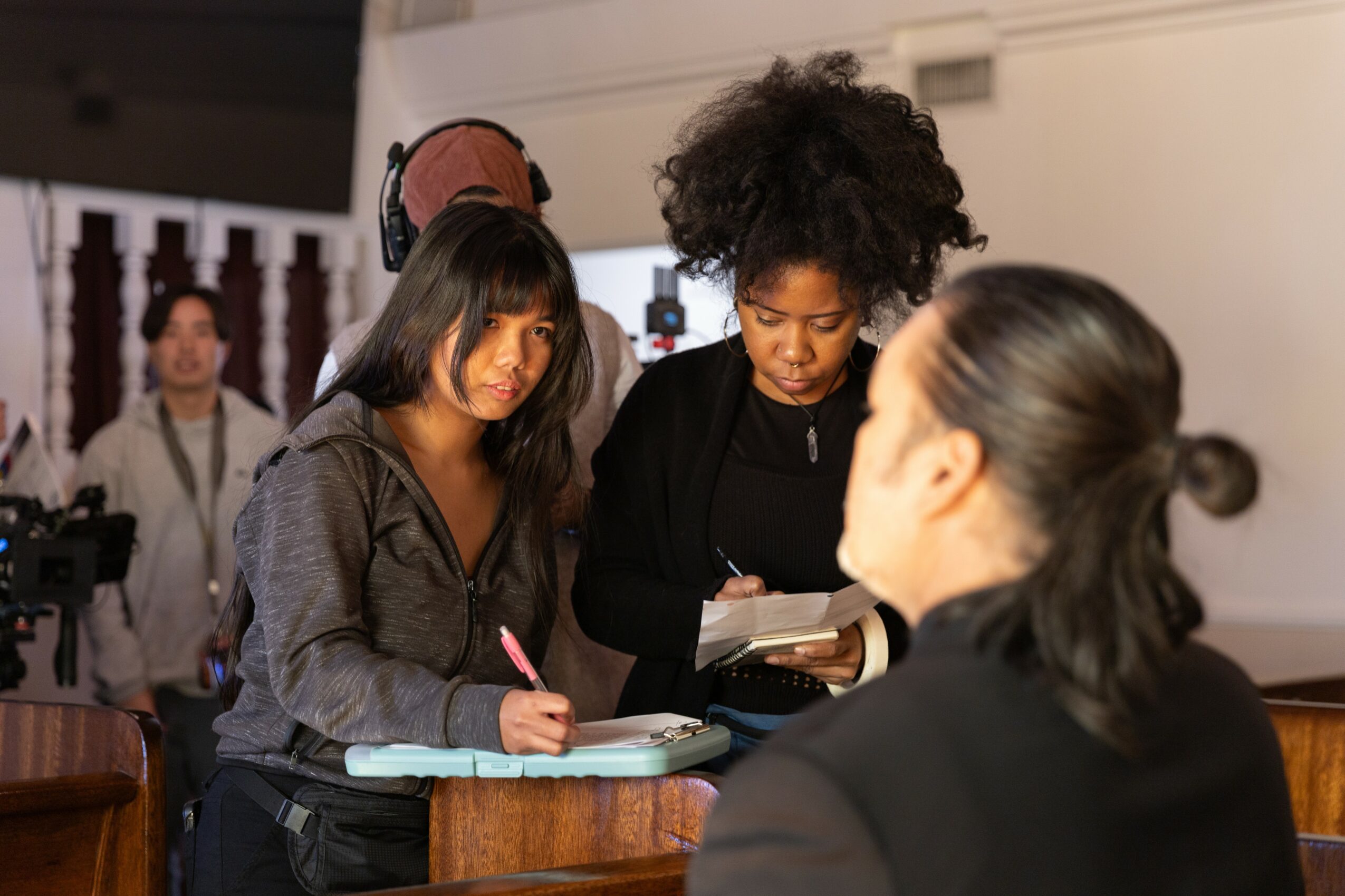 Two women and a person with a ponytail in a black suit are engaged in conversation in a room with a person in the background. 20 words.