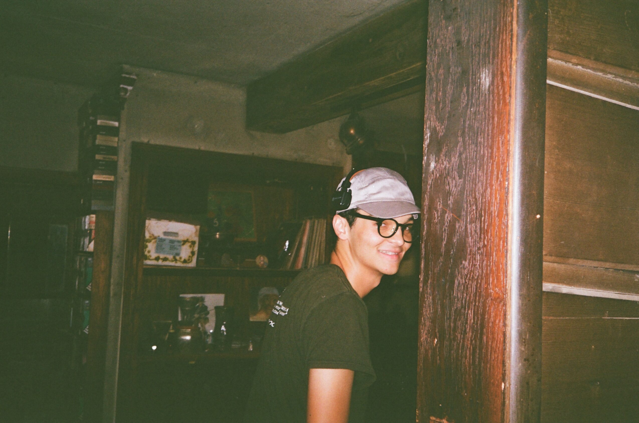 Person wearing glasses and a cap, smiling, standing indoors near a wooden pillar.