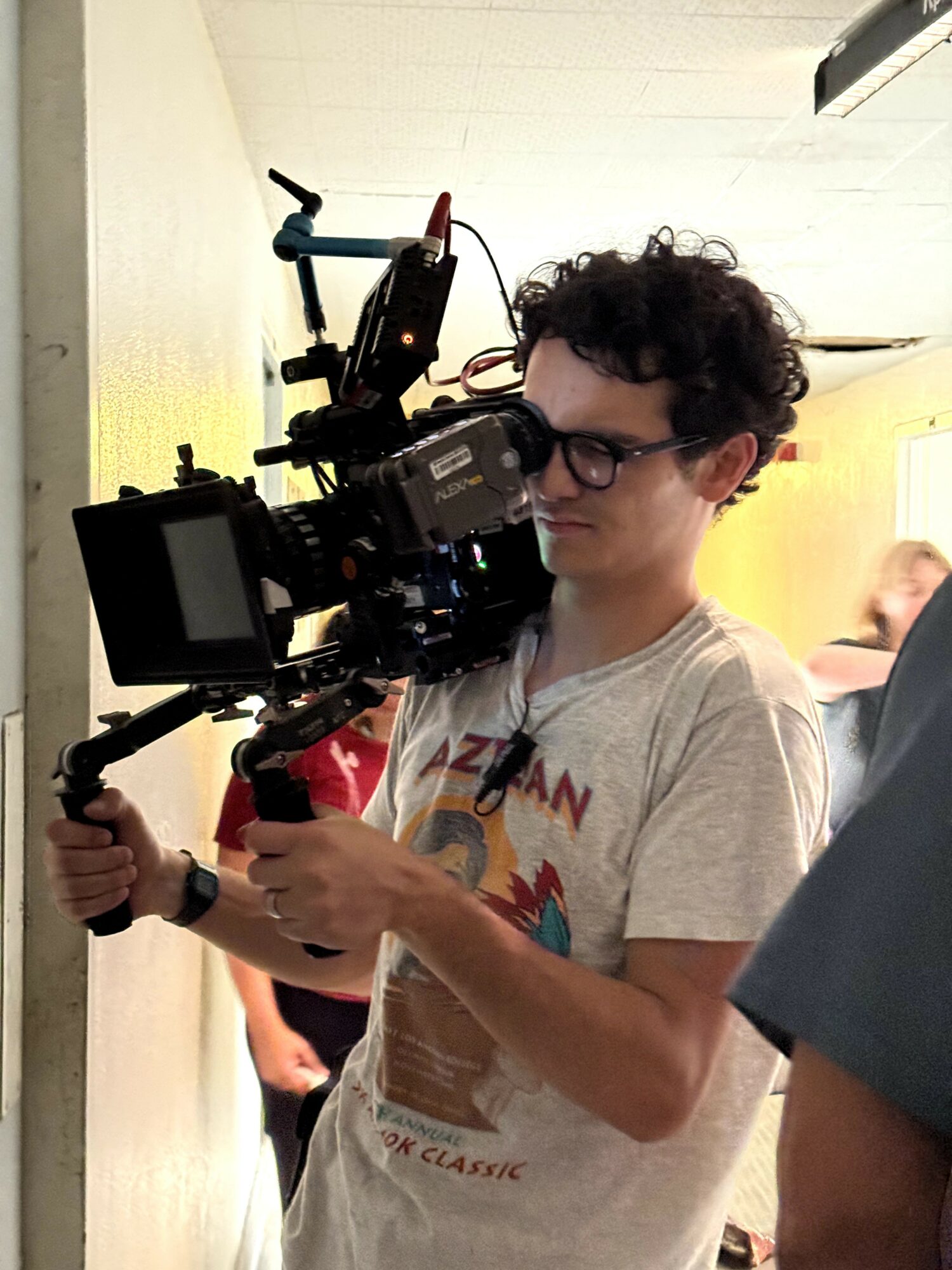 Young man with curly hair and glasses operating a professional camera, indoors with yellow and white walls.