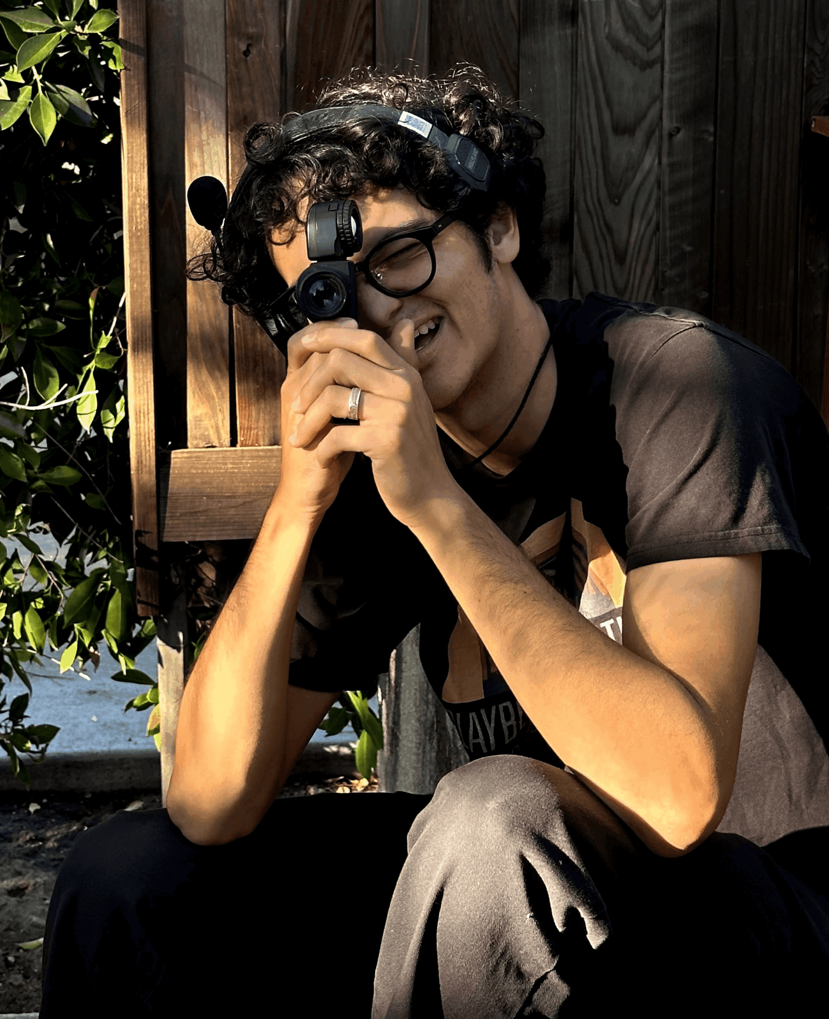 Person with curly hair and glasses smiling, holding a camera or binoculars, sitting outdoors near a wooden fence.