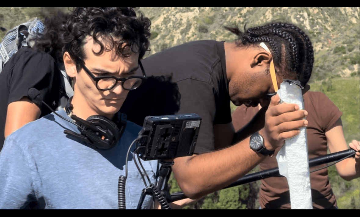 Two people outdoors, one with glasses and headphones, the other with braided hair, examining equipment with a scenic background.