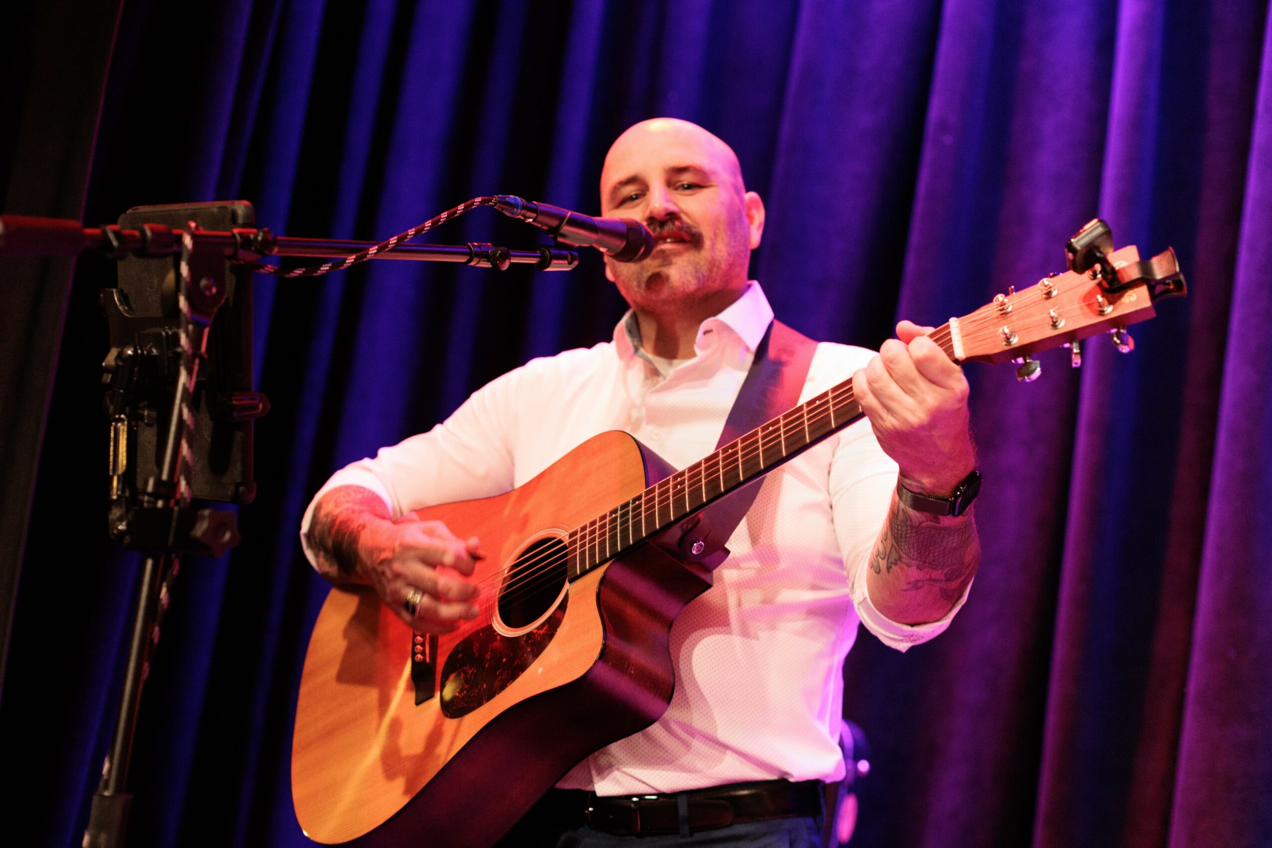 Man playing guitar and singing into microphone on stage with purple curtains behind him.