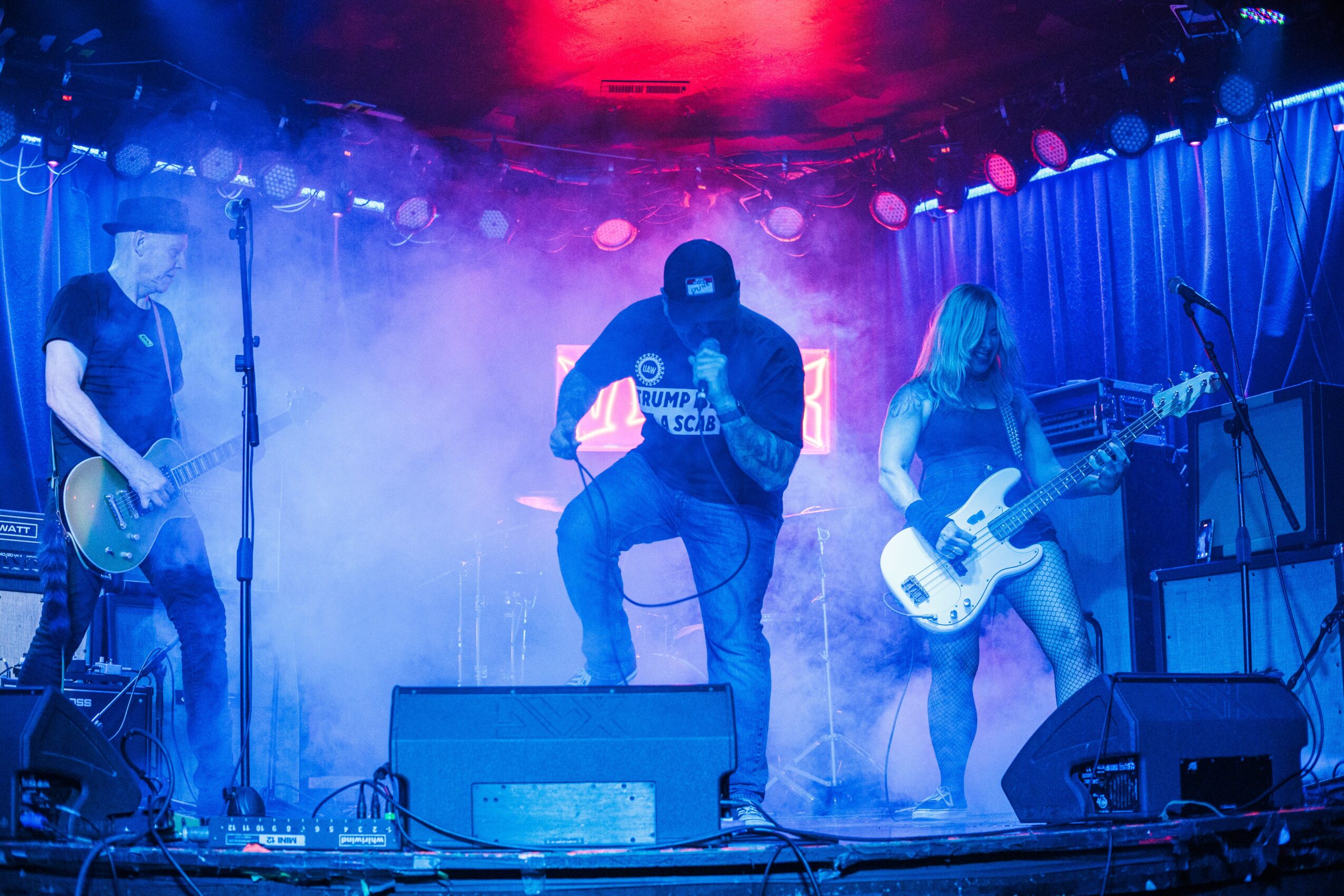 Four musicians perform on stage with colorful lighting and fog, playing guitars and singing, in a live concert setting.
