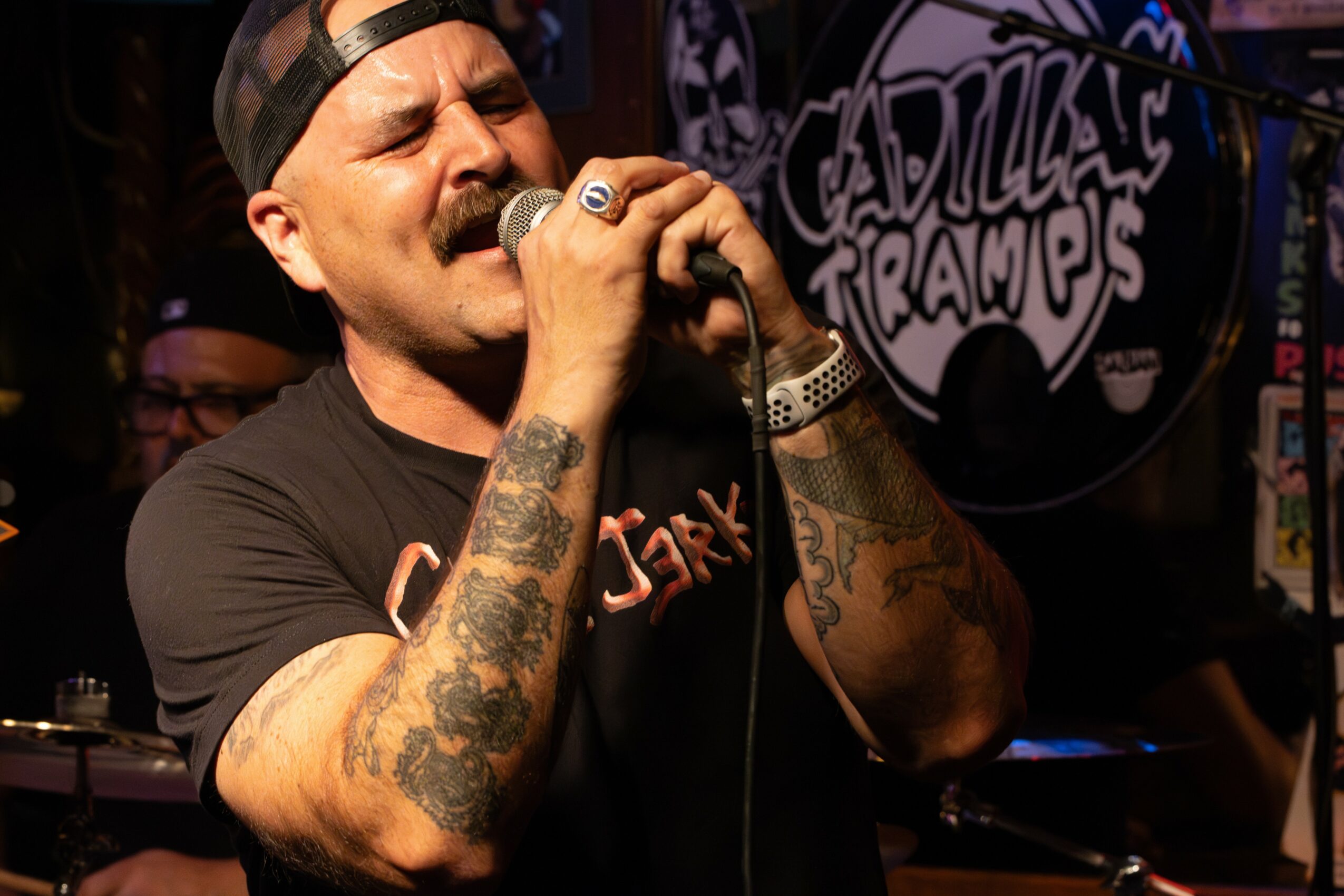 Person singing into a microphone with tattoos on arms, wearing a black cap and shirt, in a performance setting.