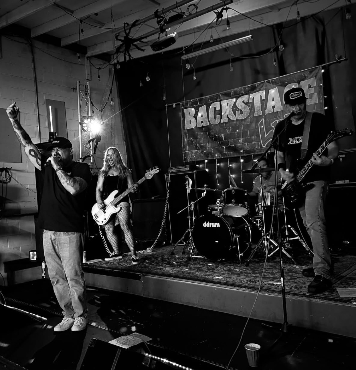 Band performs on stage with a singer, guitarist, bassist, and drummer, in a venue with a banner reading 'BACKSTA'.